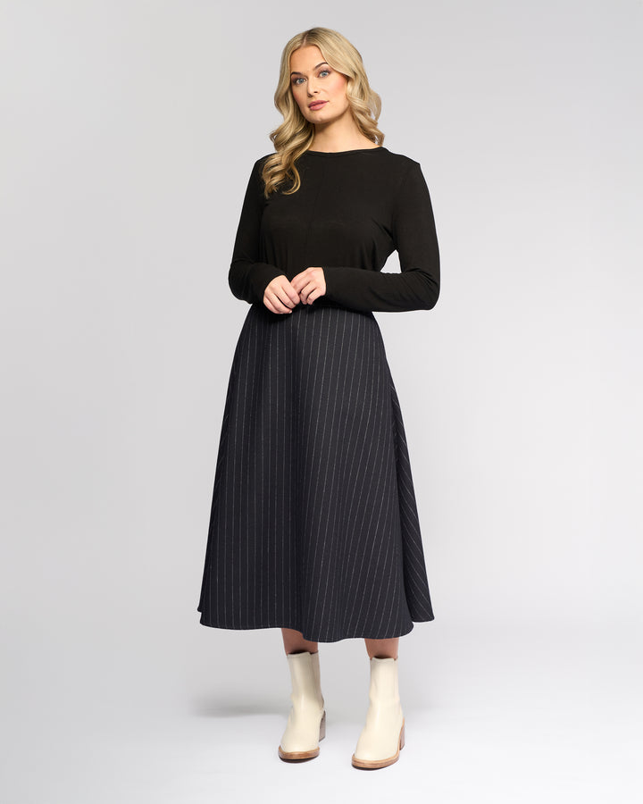 A woman with long blonde hair wears a black long-sleeve top, light ankle boots, and the Vassalli Gravity Skirt - Ink Stripe, standing against a plain light gray background.