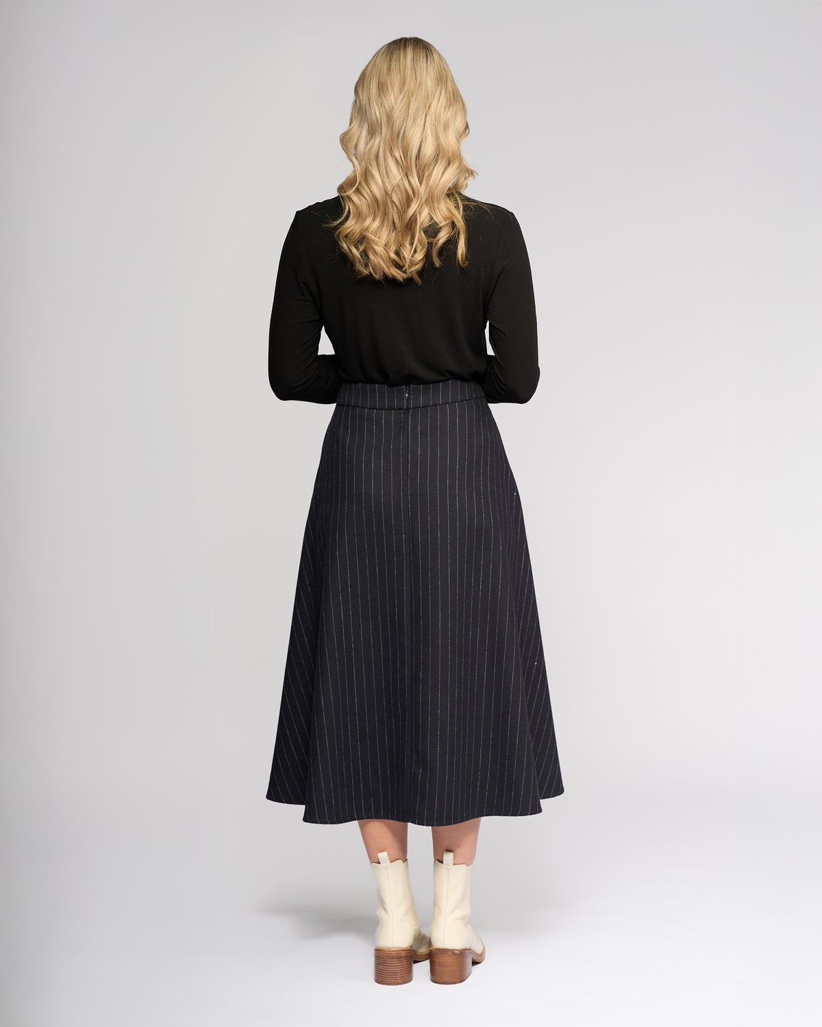 A woman with long blonde hair stands facing away, wearing a black long-sleeve top, Vassalli's Gravity Skirt in Ink Stripe, and cream ankle boots with wooden heels against a light gray background.