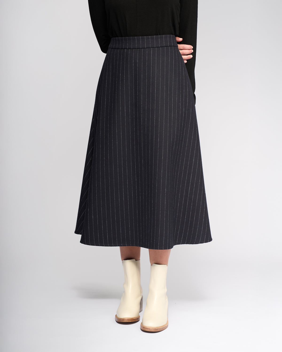 A person stands against a plain light background, arms crossed behind their back, wearing a black long-sleeve top, cream ankle boots, and the Vassalli Gravity Skirt - Ink Stripe, a black wool-blend pinstripe skirt with white lines.