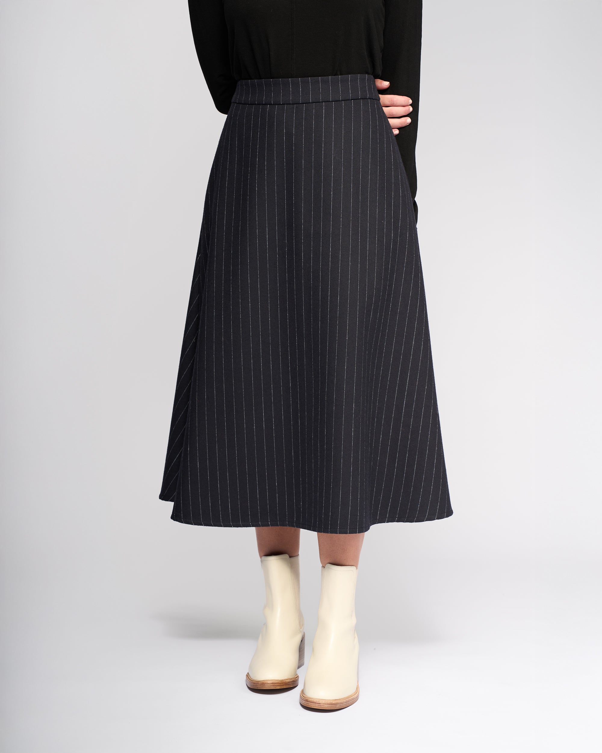 A person stands against a plain light background, arms crossed behind their back, wearing a black long-sleeve top, cream ankle boots, and the Vassalli Gravity Skirt - Ink Stripe, a black wool-blend pinstripe skirt with white lines.