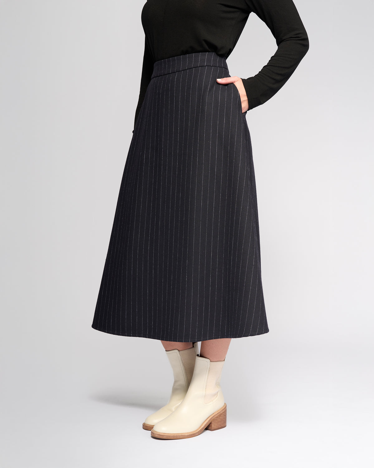A person wearing a black long-sleeve top, the Vassalli Gravity Skirt - Ink Stripe, and off-white ankle boots with wooden heels stands with one hand in their pocket against a plain background.