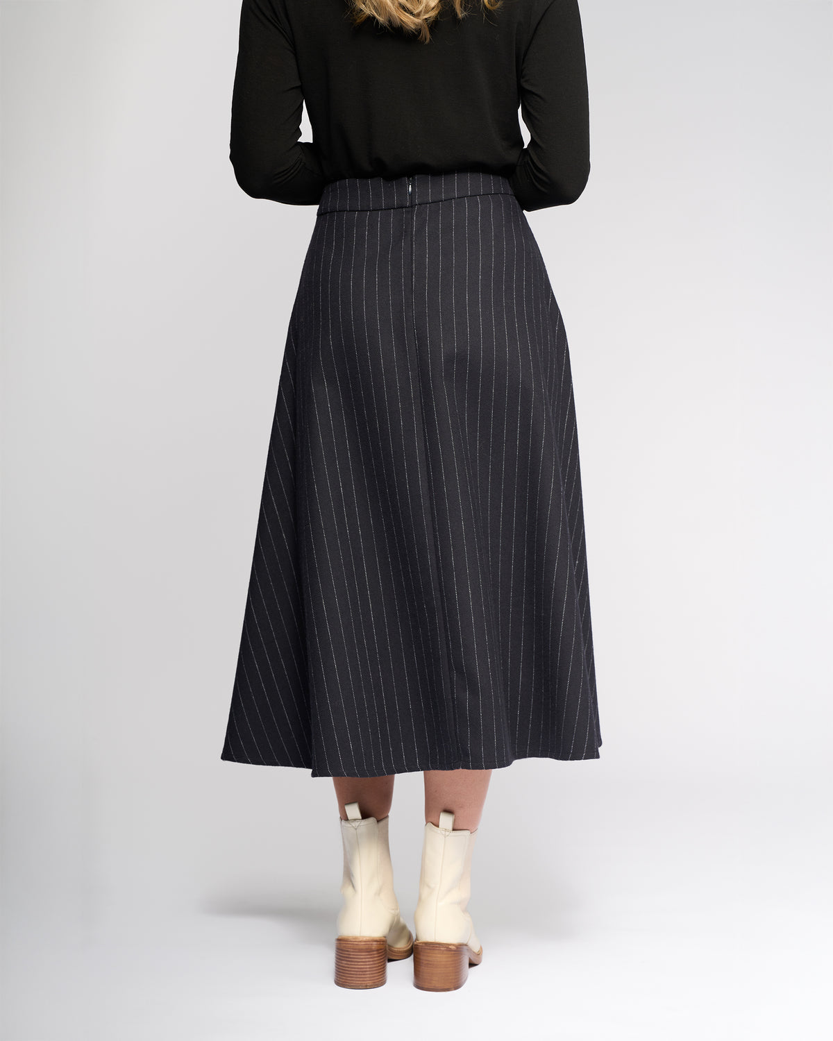 A woman stands facing away, wearing Vassalli’s Gravity Skirt - Ink Stripe, a black long-sleeve top, and cream ankle boots with wooden heels against a plain light background.