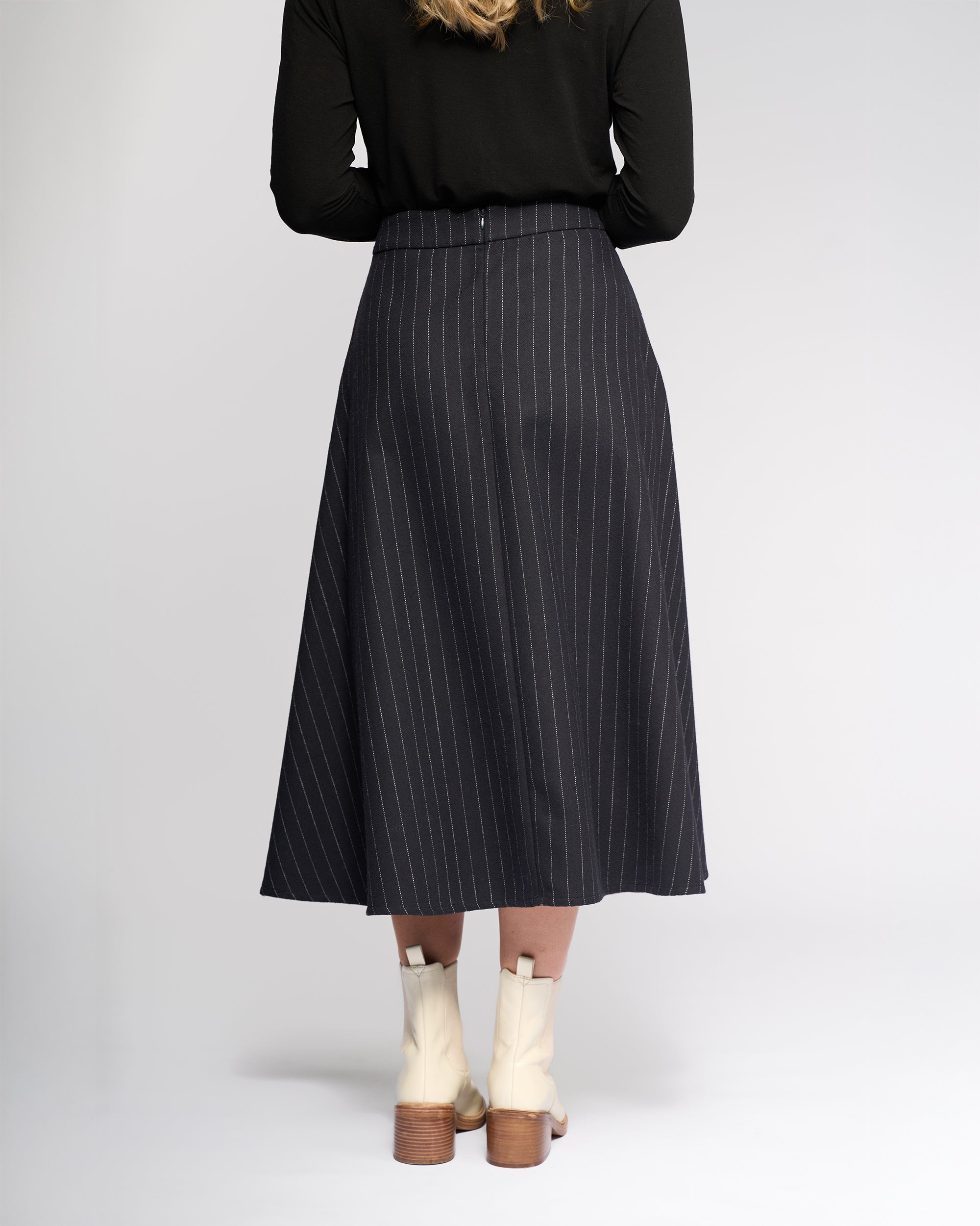 A person, seen from behind, wears a black long-sleeve top, Vassalli’s Gravity Skirt in Ink Stripe, and cream boots with wooden heels, standing against a plain light background.
