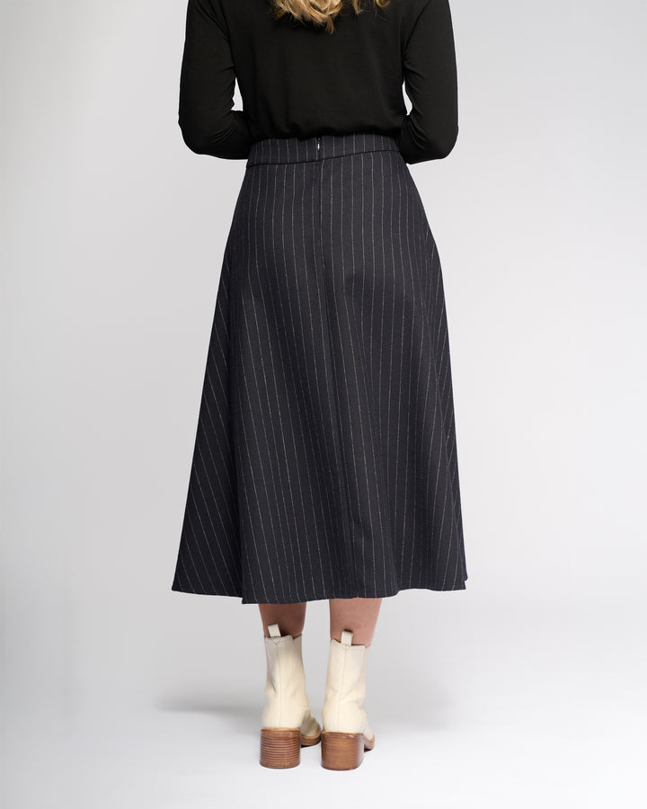 A person, seen from behind, wears a black long-sleeve top, Vassalli’s Gravity Skirt in Ink Stripe, and cream boots with wooden heels, standing against a plain light background.