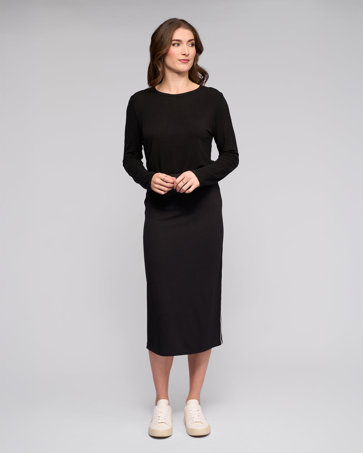 A woman stands against a light gray background, wearing the Vassalli Maple Skirt in black with white sneakers. Her wavy brown hair frames her soft smile as she looks to her left—an effortless, stylish choice for travel.