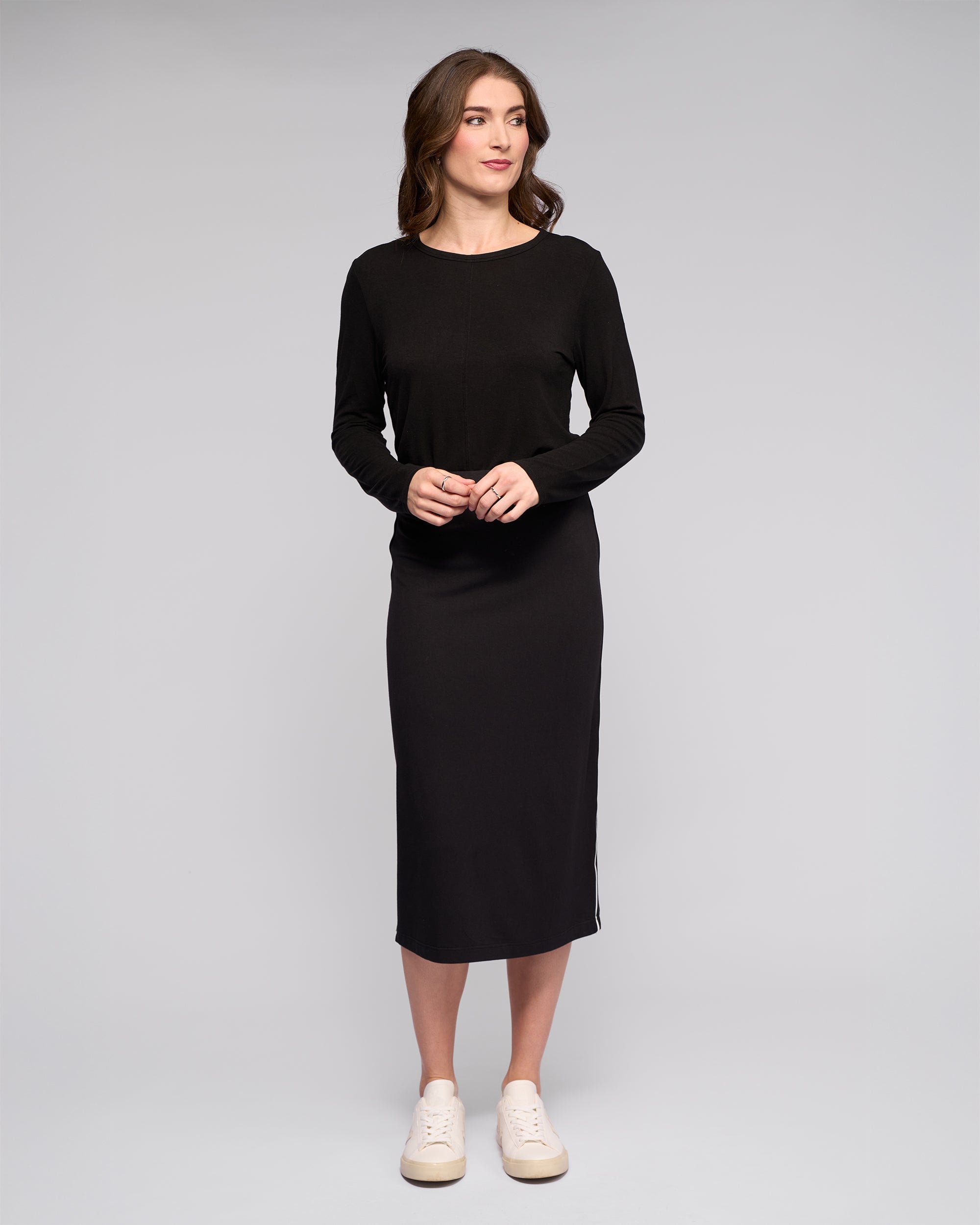A woman stands against a light gray background, wearing the Vassalli Maple Skirt in black with white sneakers. Her wavy brown hair frames her soft smile as she looks to her left—an effortless, stylish choice for travel.