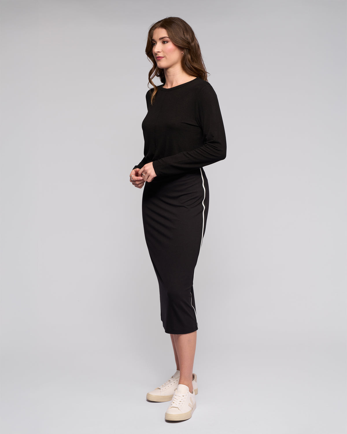 A woman with wavy brown hair stands sideways against a plain background, wearing Vassalli’s Maple Skirt – Black, featuring white side stripes and a stretchy midi fit. She pairs it with a long-sleeved black top and white sneakers, hands together in front.