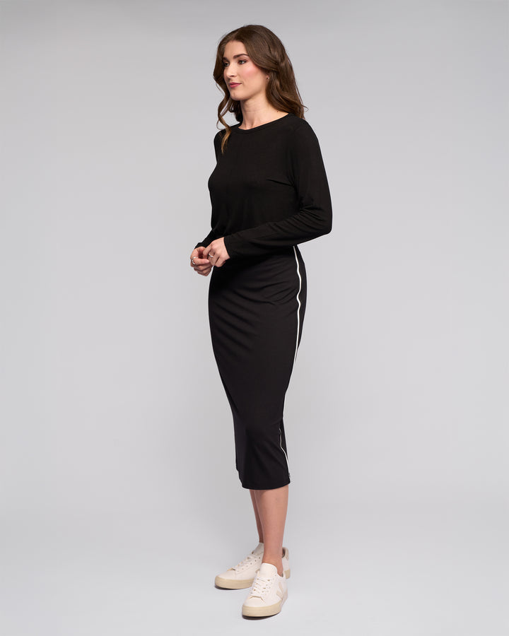 A woman with wavy brown hair stands sideways against a plain background, wearing Vassalli’s Maple Skirt – Black, featuring white side stripes and a stretchy midi fit. She pairs it with a long-sleeved black top and white sneakers, hands together in front.