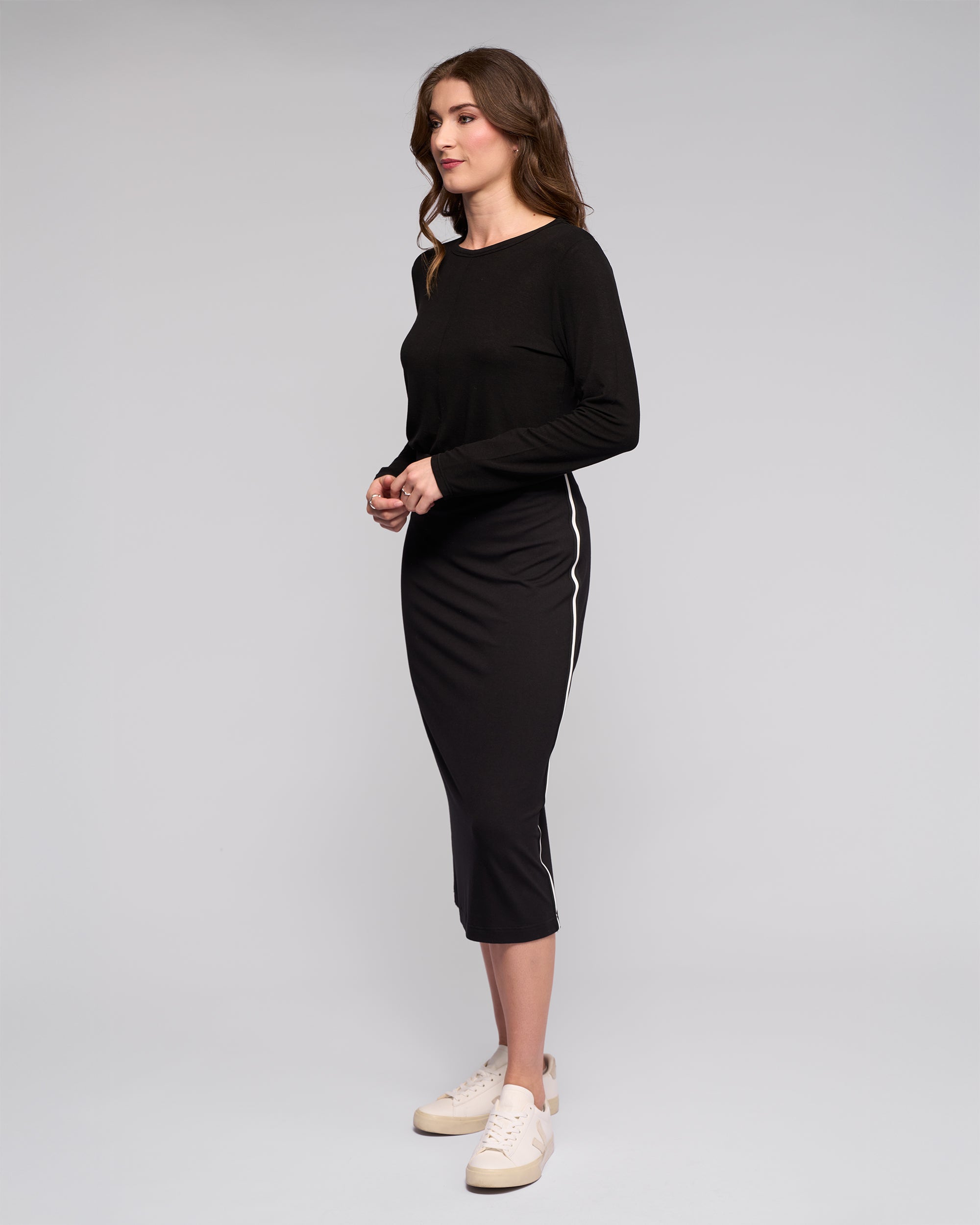 A woman stands in profile against a plain background, wearing a black long-sleeve top, white sneakers, and the Maple Skirt - Black by Vassalli: a travel wear, midi-length skirt with white side stripes in stretchy fabric.
