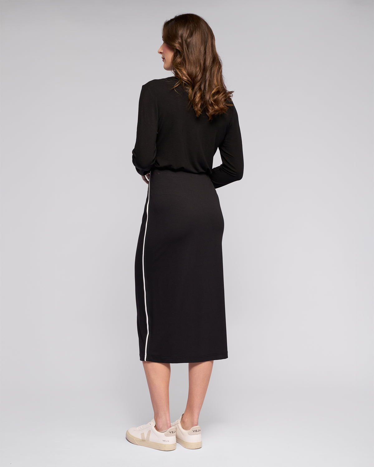 A woman with wavy brown hair faces away, wearing the Vassalli Maple Skirt in black—a long-sleeved, stretchy midi skirt with a white side stripe—paired with beige platform sneakers against a plain light gray background.