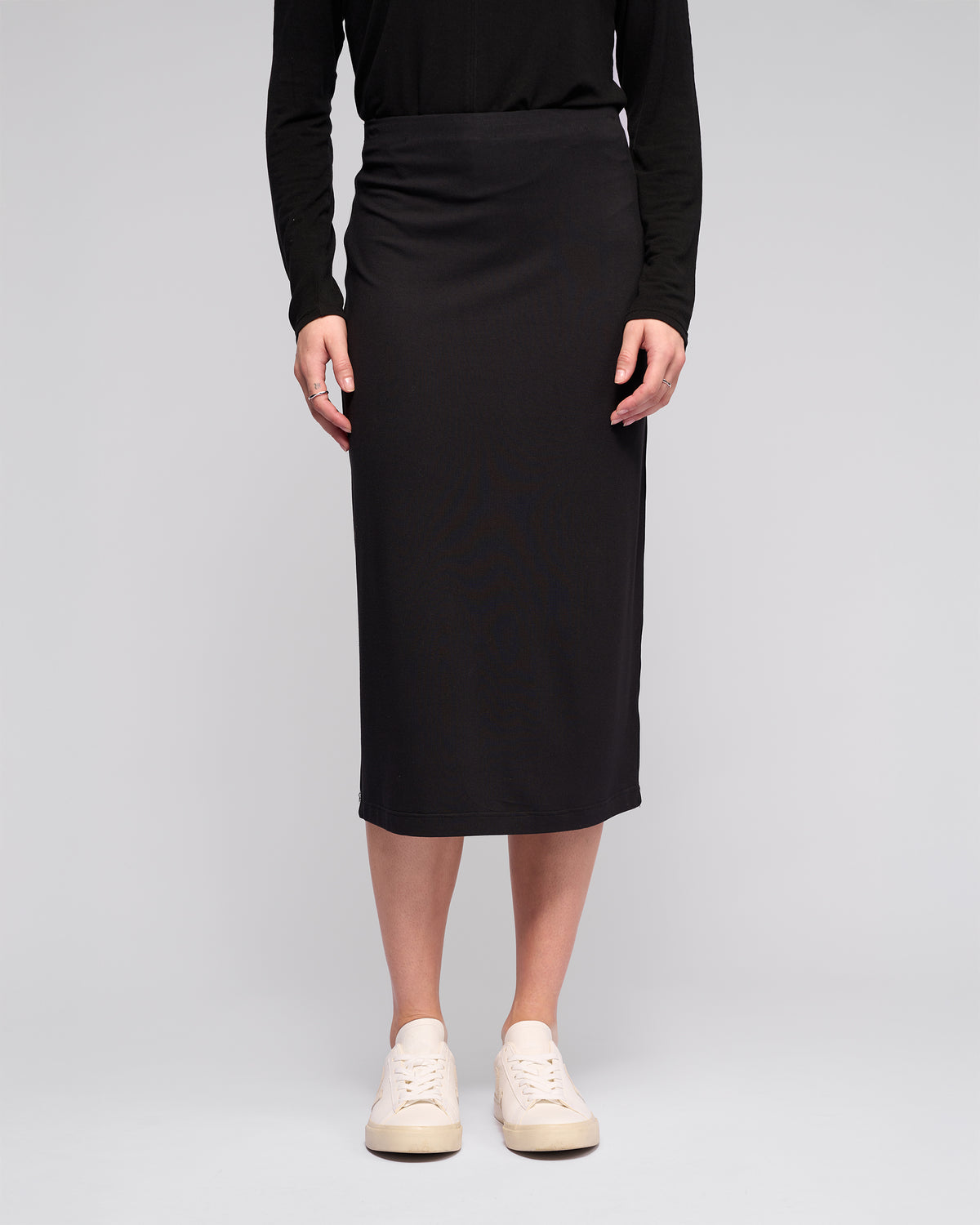 A person showcases the Vassalli Maple Skirt in black, paired with a black long-sleeve top and white sneakers against a plain light gray background. The photo is cropped from the shoulders down.