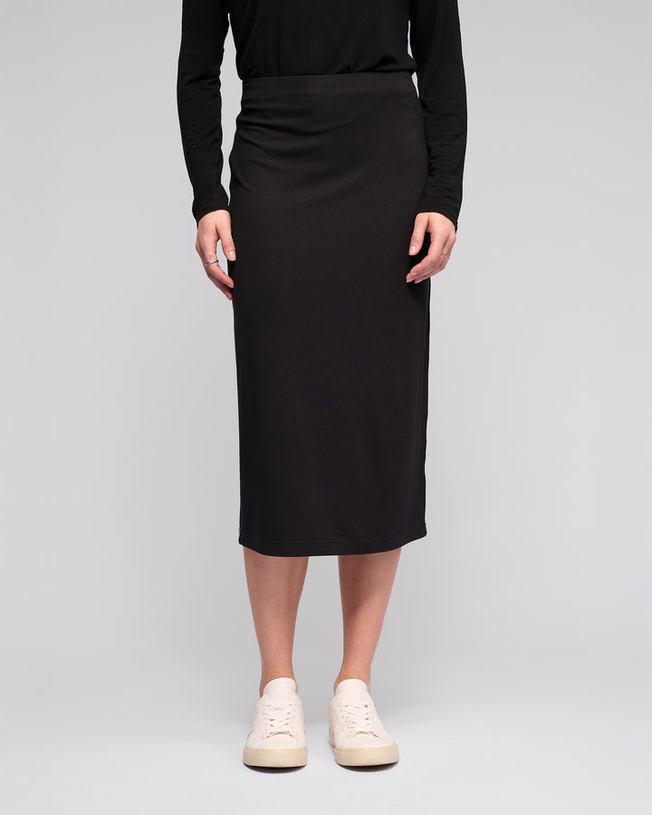 A person models the Vassalli Maple Skirt - Black, paired with a black long-sleeve top and white sneakers, against a plain light gray background. The upper body is partially out of frame. Perfect for stylish travel wear.