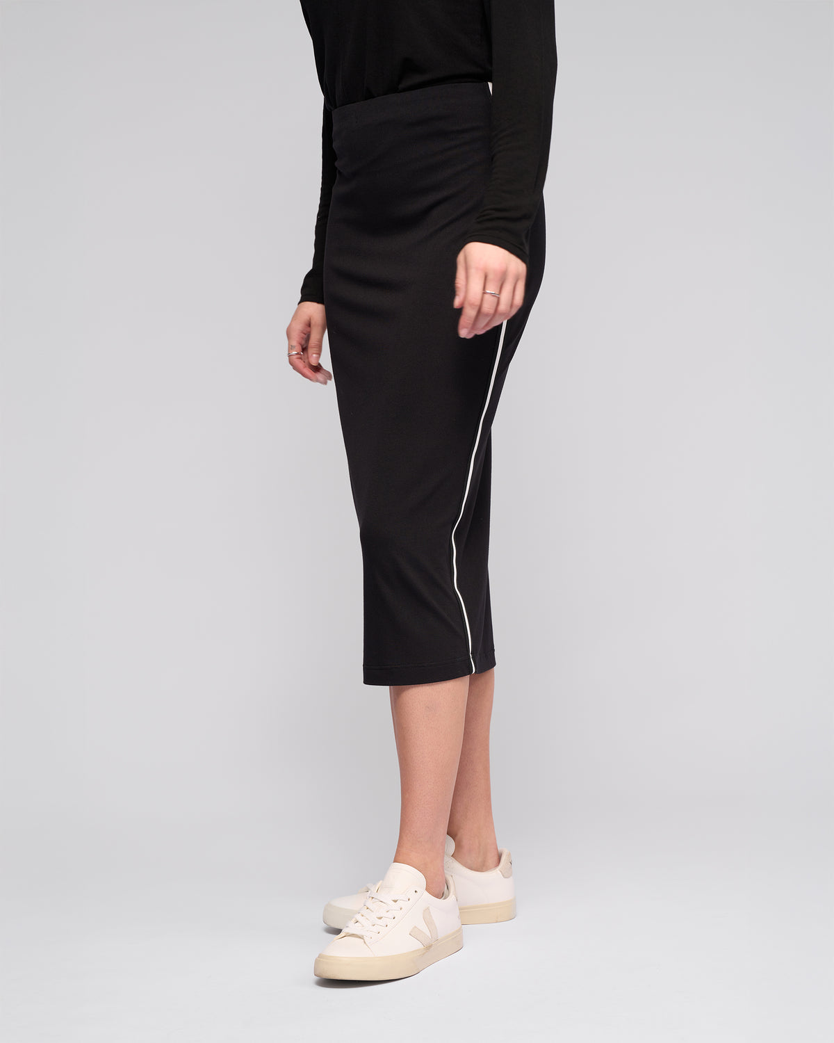 A person wears the Vassalli Maple Skirt – Black, a stretchy, midi-length pencil skirt with a white side stripe, paired with a black long-sleeved top and white sneakers against a light gray background—ideal for stylish travel.