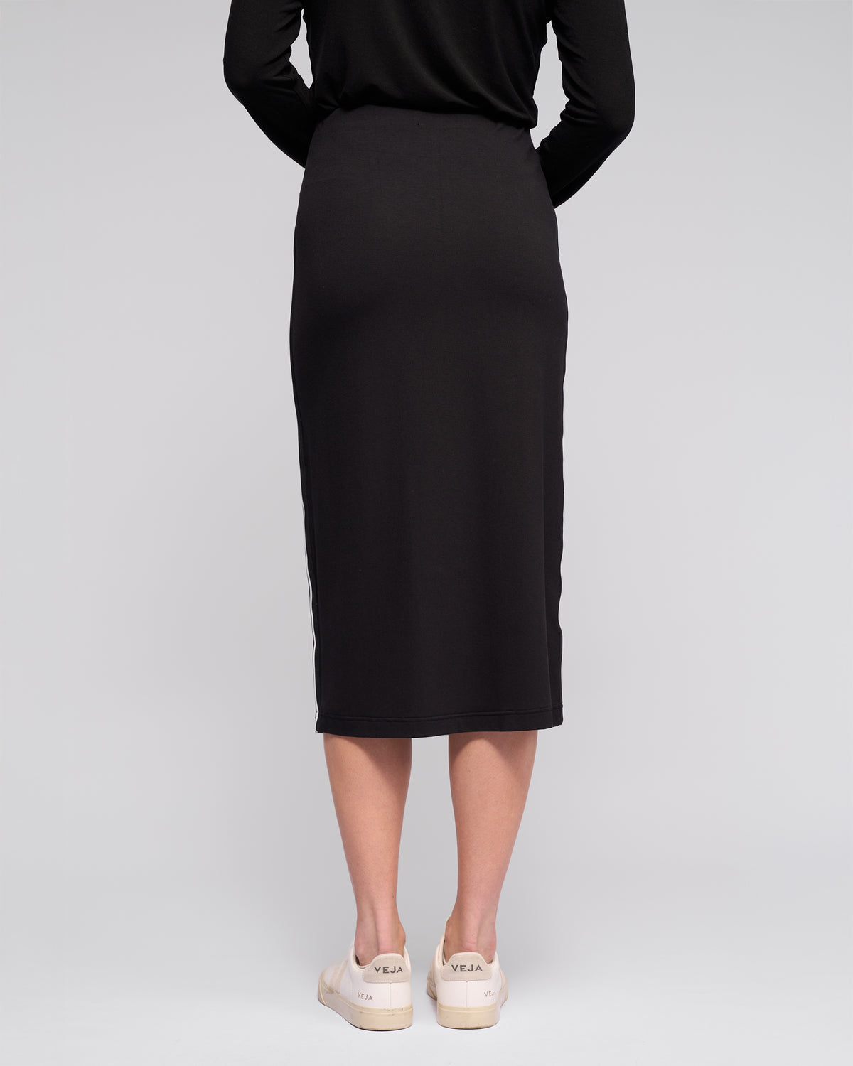 A person stands against a light grey background, seen from the back, wearing the Vassalli Maple Skirt in black—midi-length and stretchy—paired with off-white sneakers for comfortable travel style.