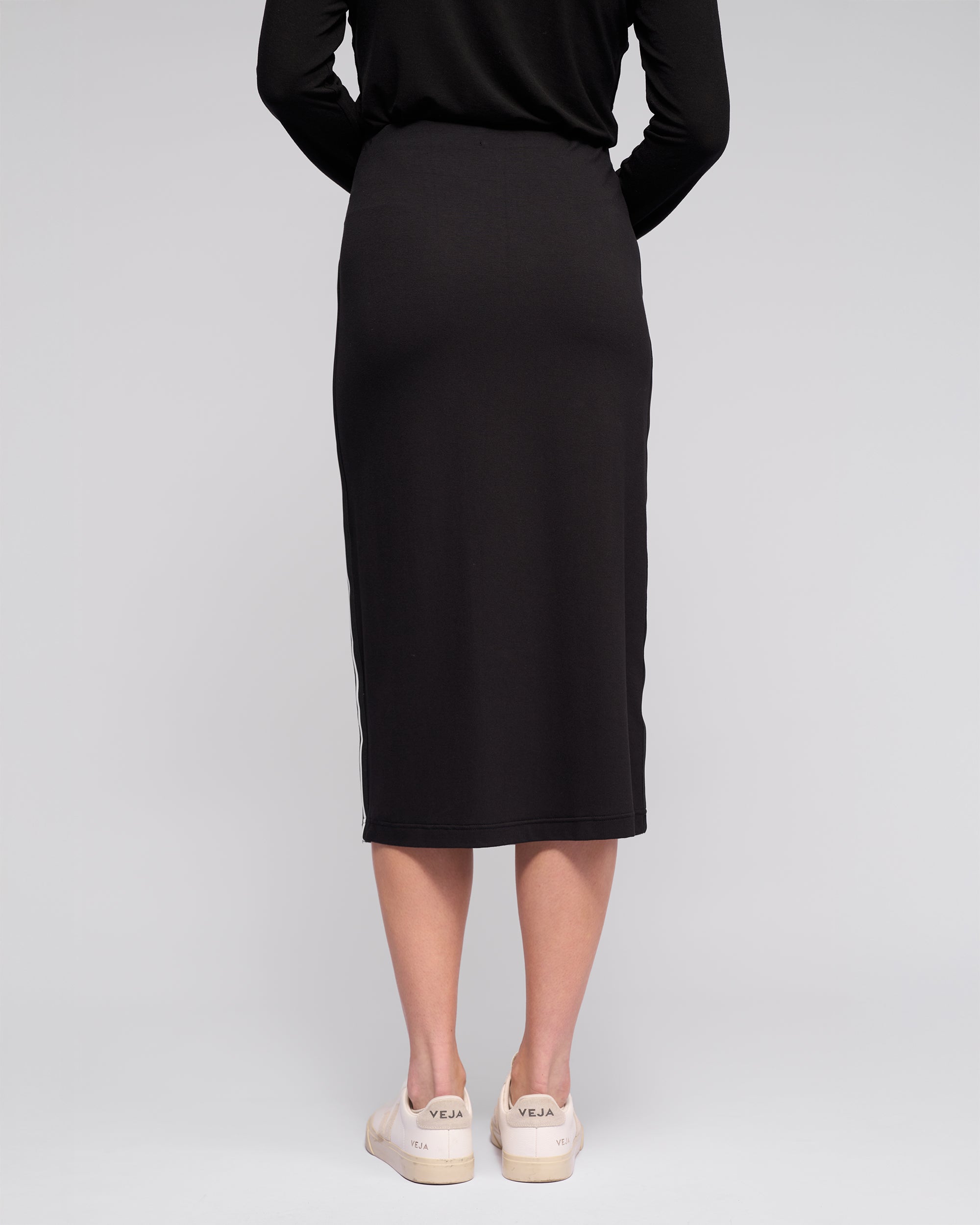 A person stands against a light grey background, seen from the back, wearing the Vassalli Maple Skirt in black—midi-length and stretchy—paired with off-white sneakers for comfortable travel style.