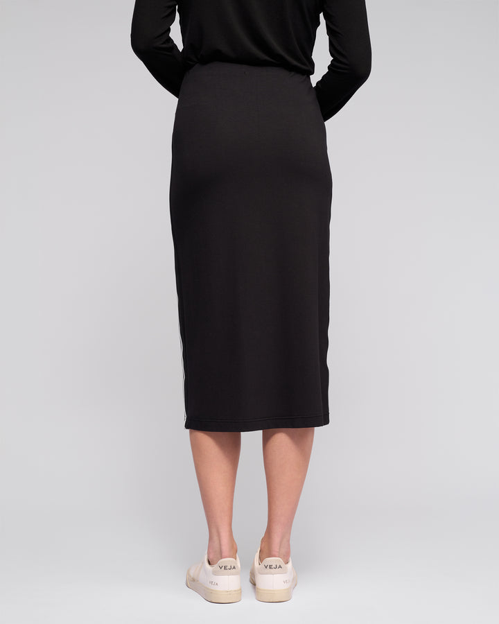 A person is seen from behind in the Vassalli Maple Skirt - Black, a stretchy midi skirt, paired with white Veja sneakers against a plain light background—an ideal stylish choice for travel.