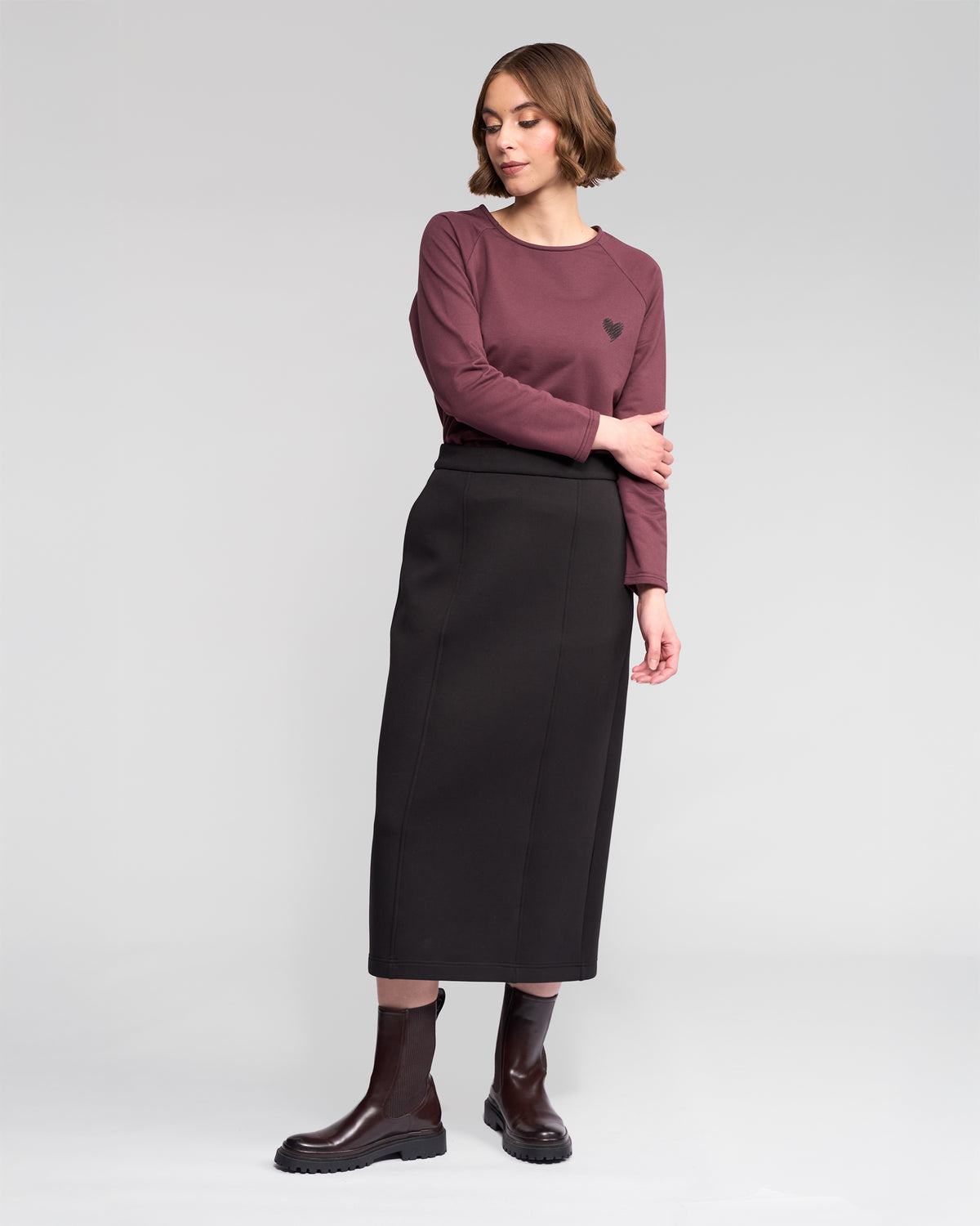 A woman stands against a light gray background wearing the Vassalli Falcon Skirt in black, paired with a mauve long-sleeve top and dark brown ankle boots. She looks to her left with a relaxed posture.