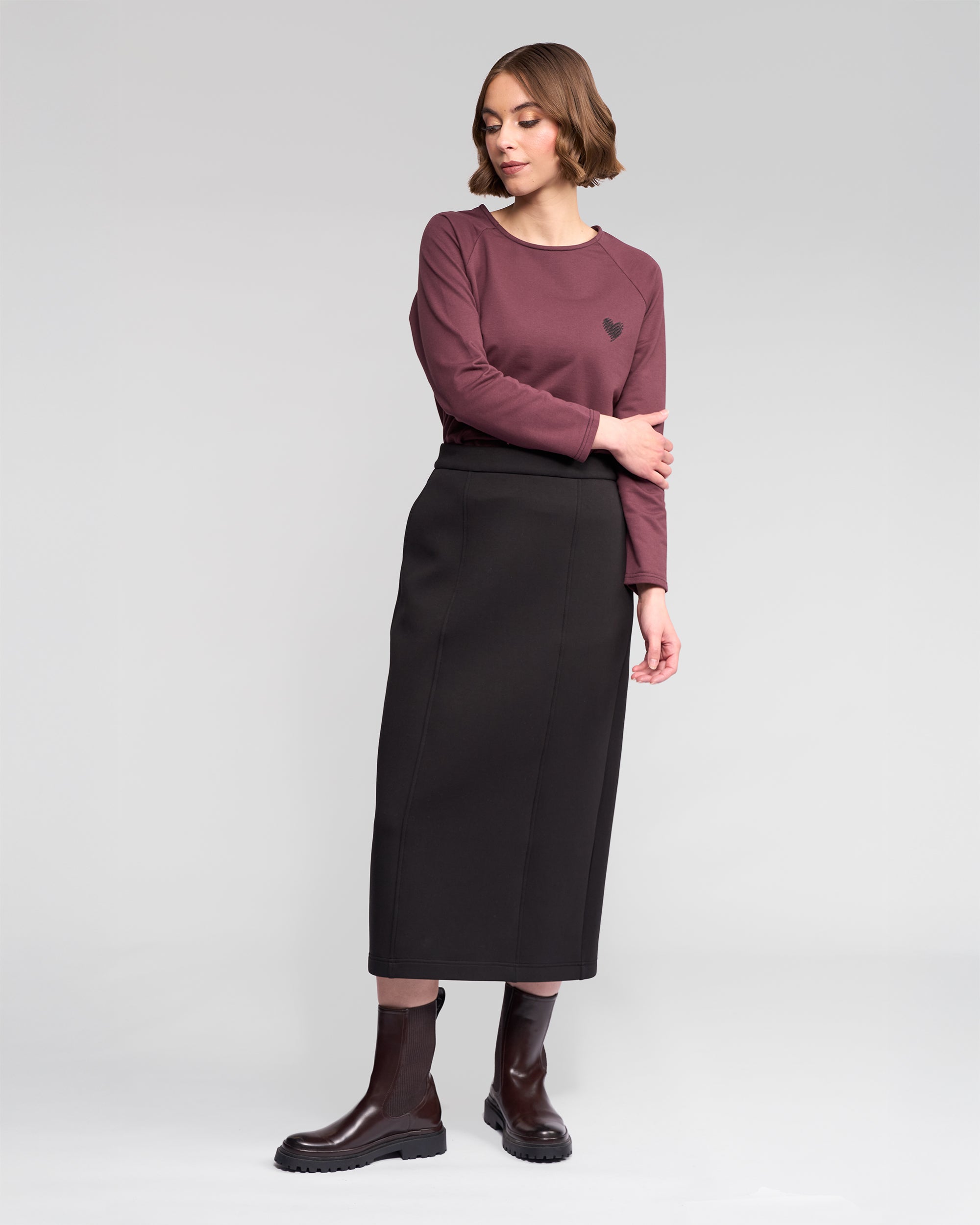 A woman stands against a light gray background wearing the Vassalli Falcon Skirt in black, paired with a mauve long-sleeve top and dark brown ankle boots. She looks to her left with a relaxed posture.