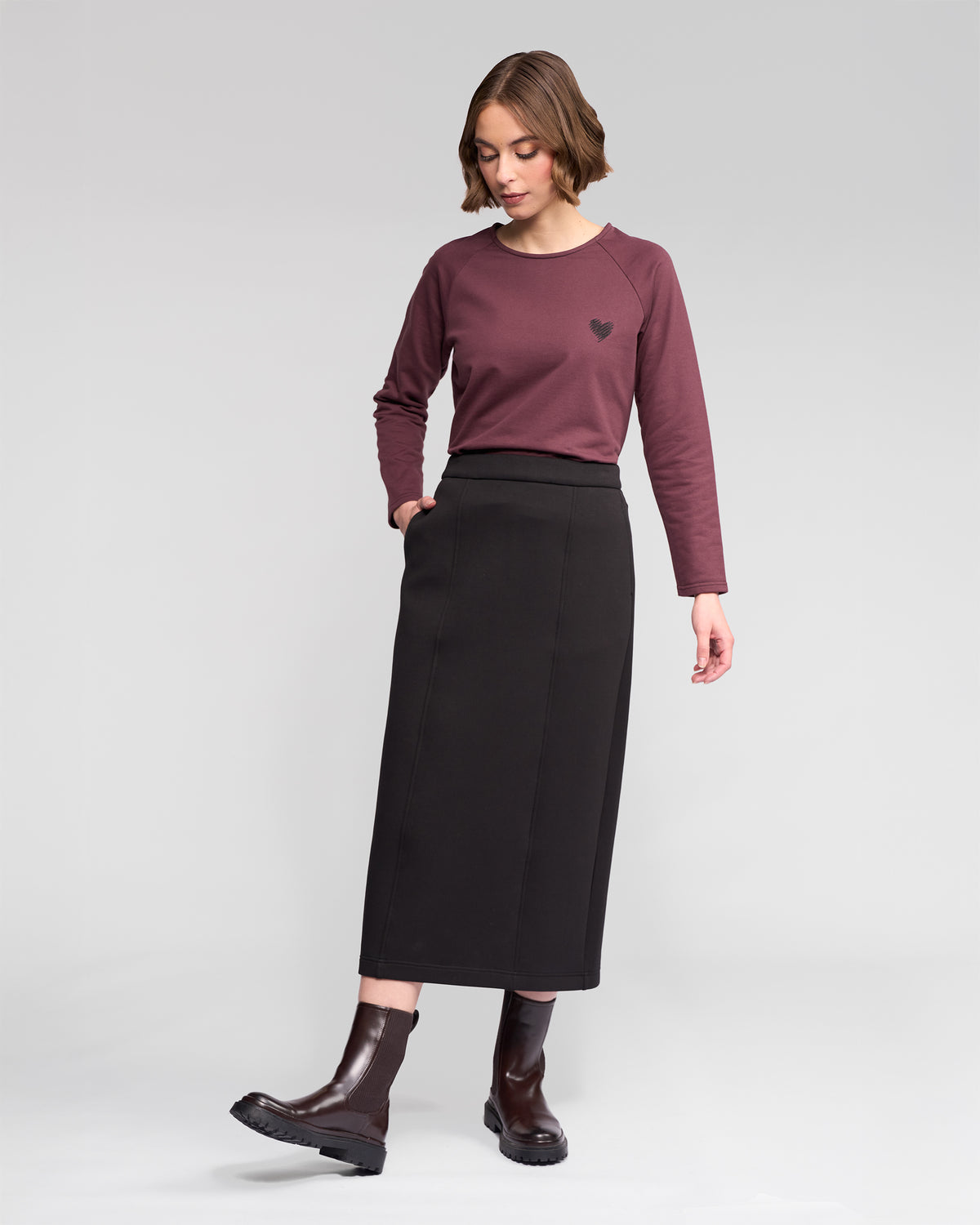 A woman stands against a plain light gray background, wearing the Vassalli Falcon Skirt in black. She pairs it with a maroon long-sleeve top featuring a small heart design and dark brown ankle boots, with her hands tucked in her skirt pockets.