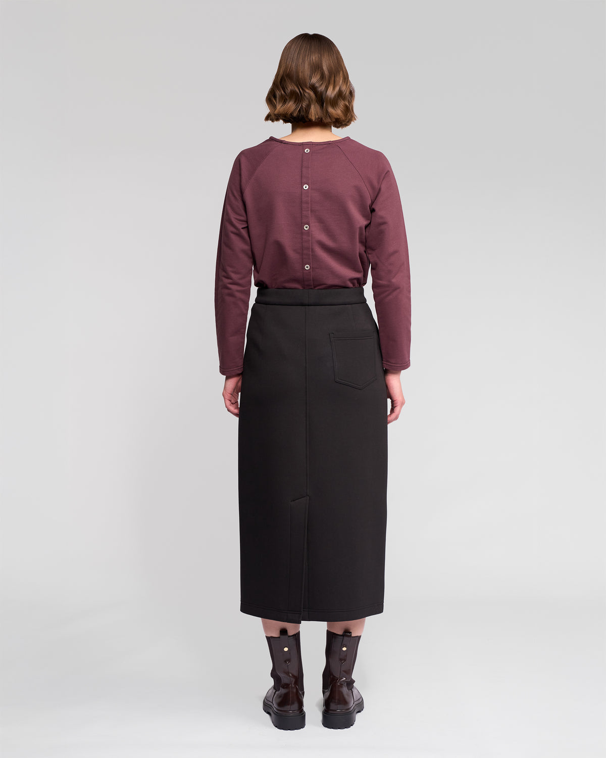 A person with short brown hair faces away, wearing a mauve long-sleeve top with back buttons, Vassalli's Falcon Skirt in black, and black ankle boots against a plain light background.