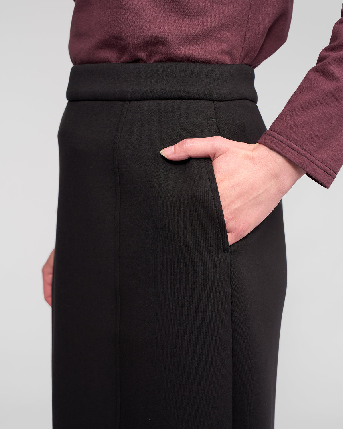 A person shown from the waist down, wearing the Vassalli Falcon Skirt in black—a high-waisted, winter-weight midi skirt—paired with a burgundy long-sleeve top, one hand in the skirt pocket, against a plain background.