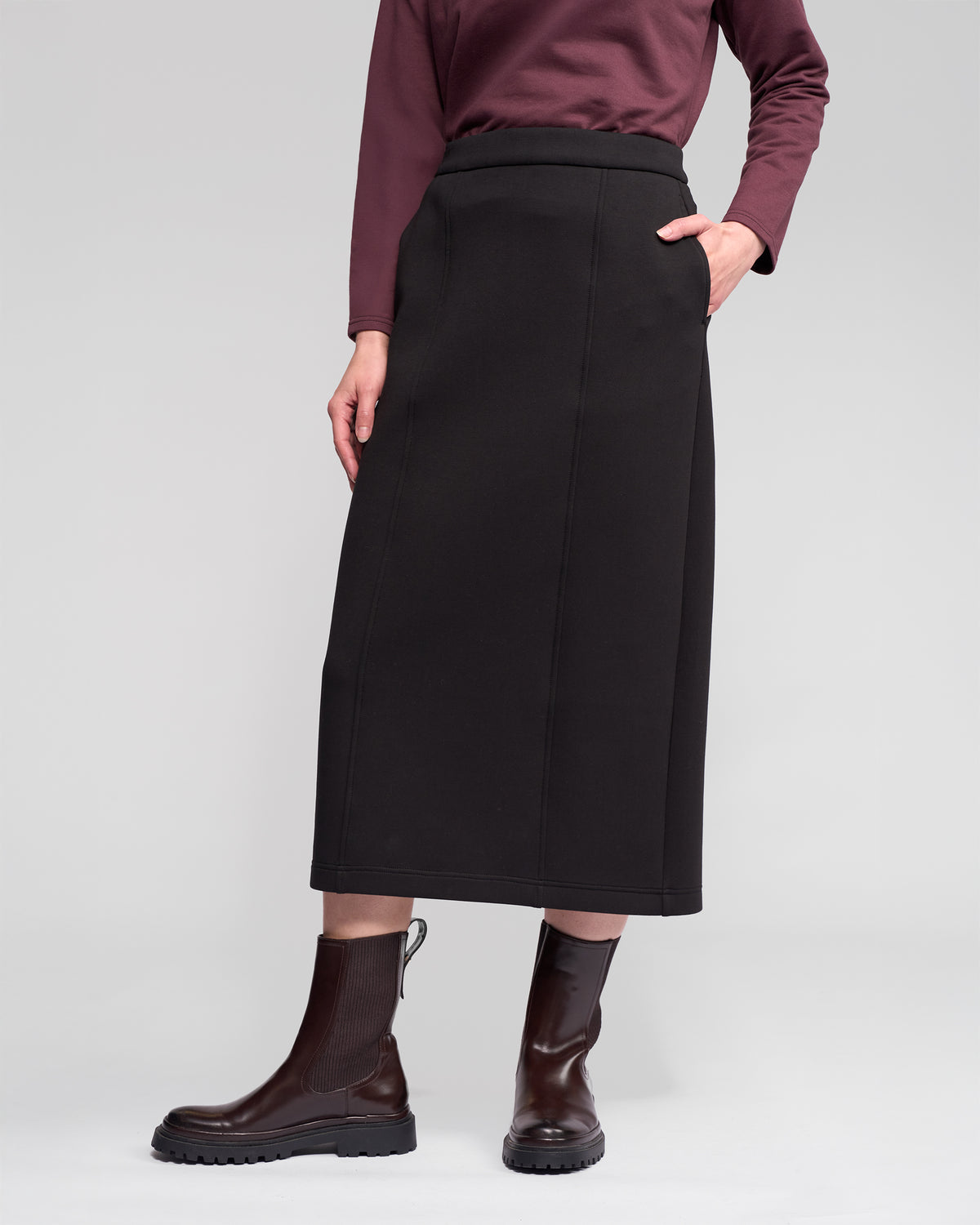 A person wears the Vassalli Falcon Skirt in black, paired with a maroon long-sleeve top and dark brown ankle boots, standing with one hand in their pocket against a plain light gray background.