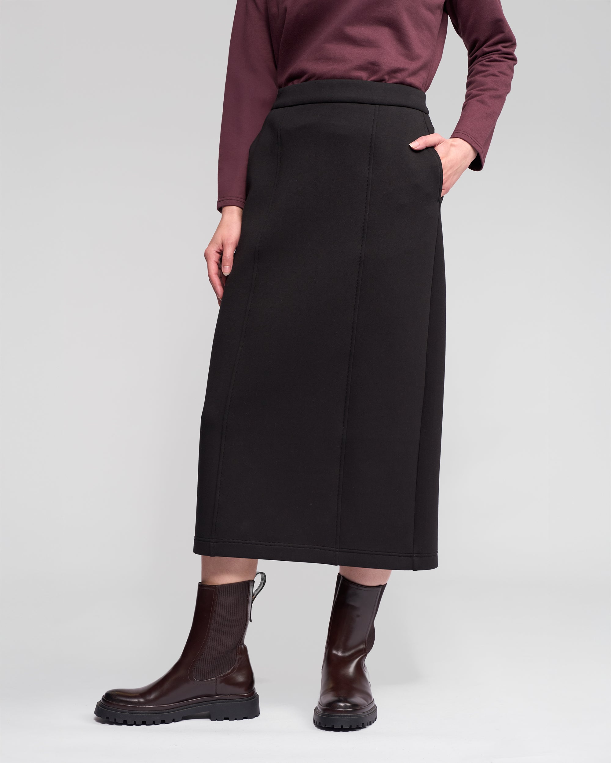 A person wears the Vassalli Falcon Skirt in black, paired with a maroon long-sleeve top and dark brown ankle boots, standing with one hand in their pocket against a plain light gray background.