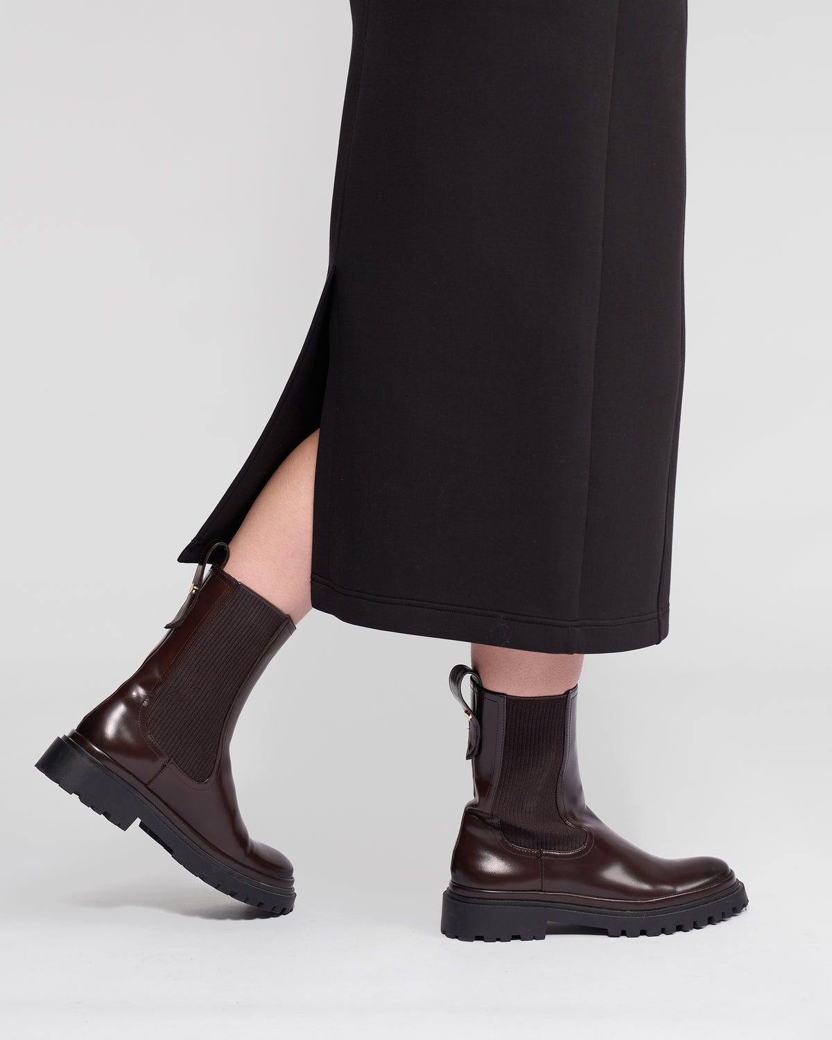 A person wears the Vassalli Falcon Skirt in black, featuring a midi length and side slit, paired with dark brown chunky ankle boots, standing against a plain light background.