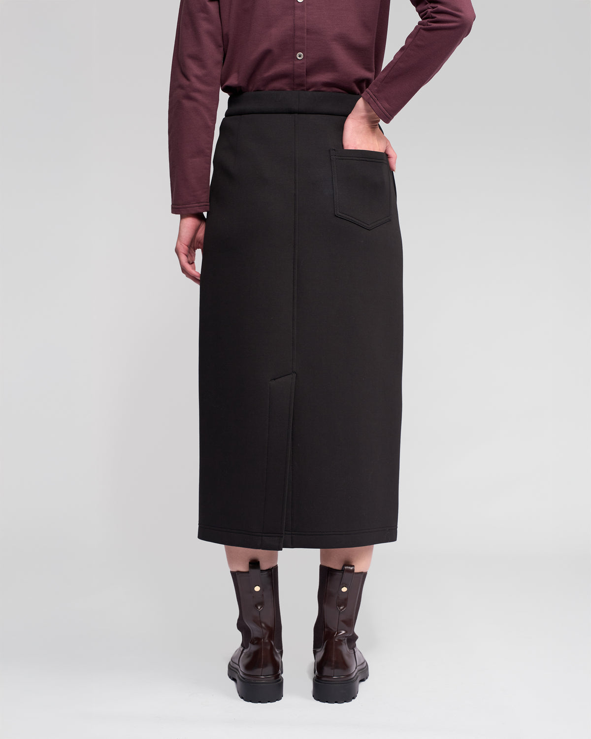 A person stands facing away, wearing a dark maroon long-sleeve shirt, Vassalli's Falcon Skirt in black with a back pocket, and dark ankle boots with buckles. One hand rests in the skirt pocket.