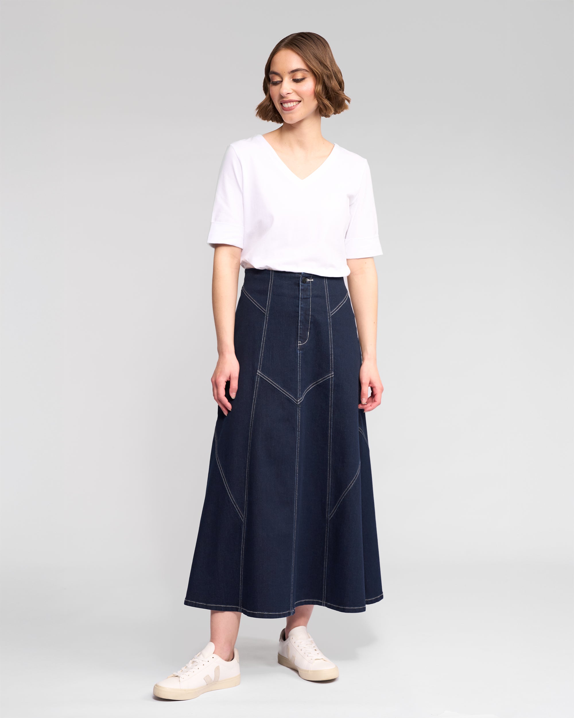 A woman smiles and looks to the side, wearing a white V-neck shirt, white sneakers, and the Vassalli Akari Skirt in ink denim with contrast white stitching against a plain background.