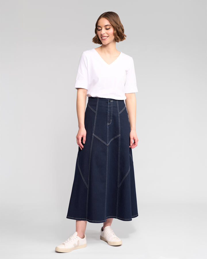 A person stands against a plain background, smiling as they wear a white V-neck top, white sneakers, and the Vassalli Akari Skirt in Ink Denim featuring contrast white stitching. They are looking slightly to the side.