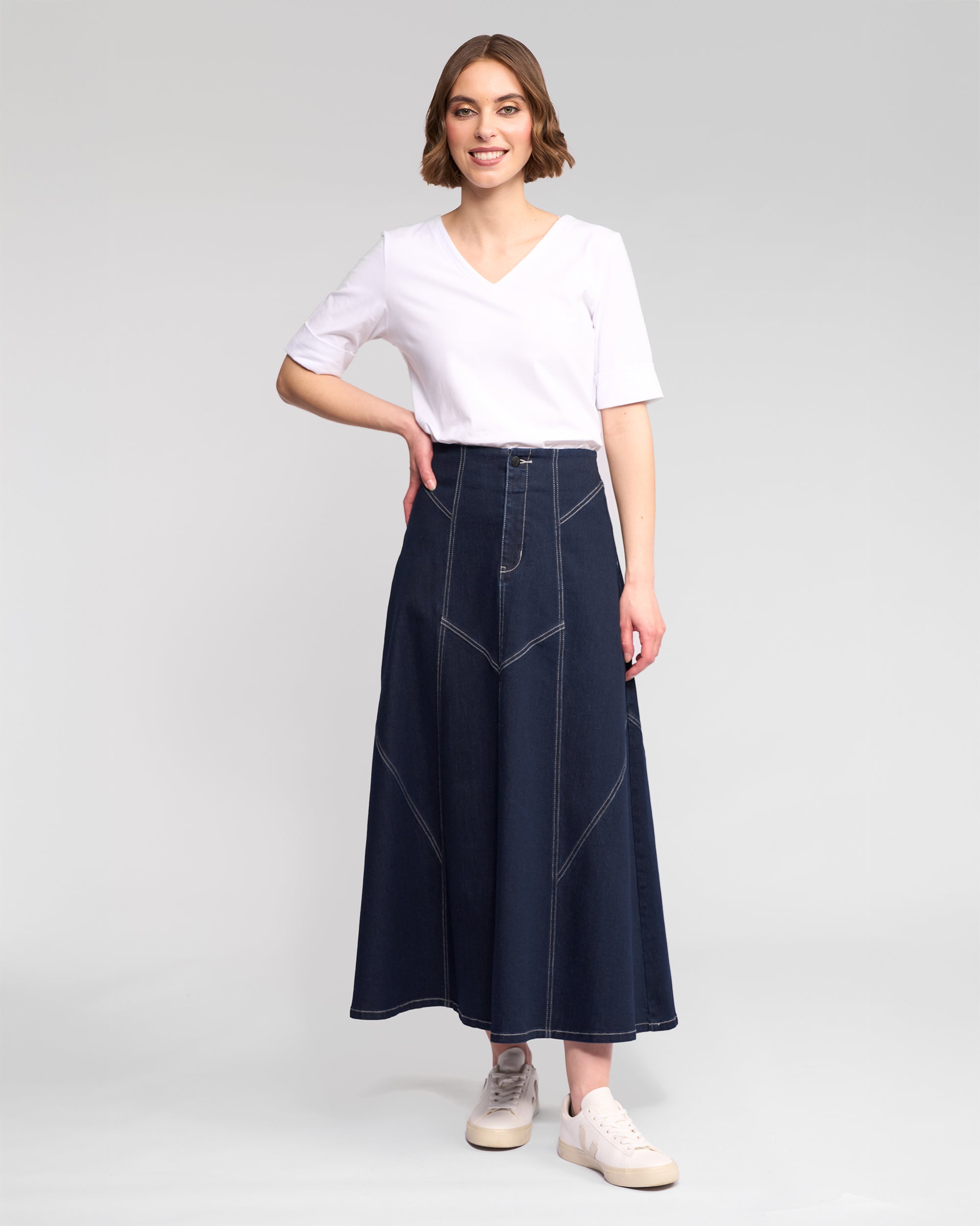 A woman smiles in a white V-neck T-shirt, white sneakers, and the Vassalli Akari Skirt in ink denim with contrast stitching, standing against a plain light gray background.