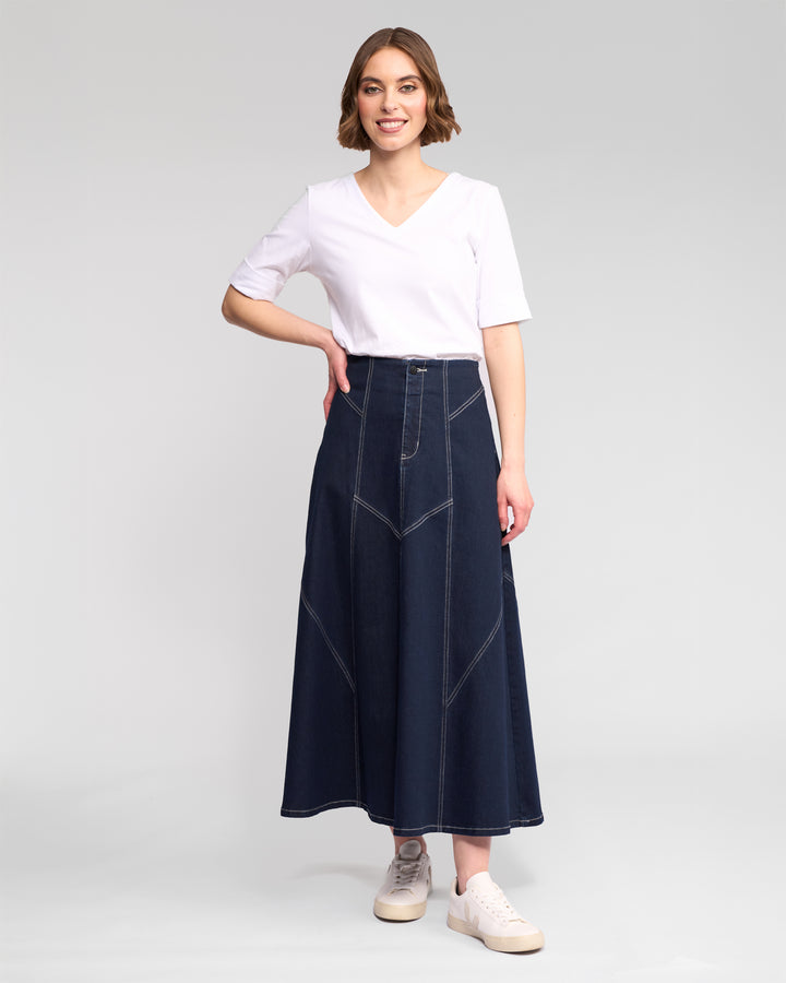 A woman smiles in a white V-neck T-shirt, white sneakers, and the Vassalli Akari Skirt in ink denim with contrast stitching, standing against a plain light gray background.
