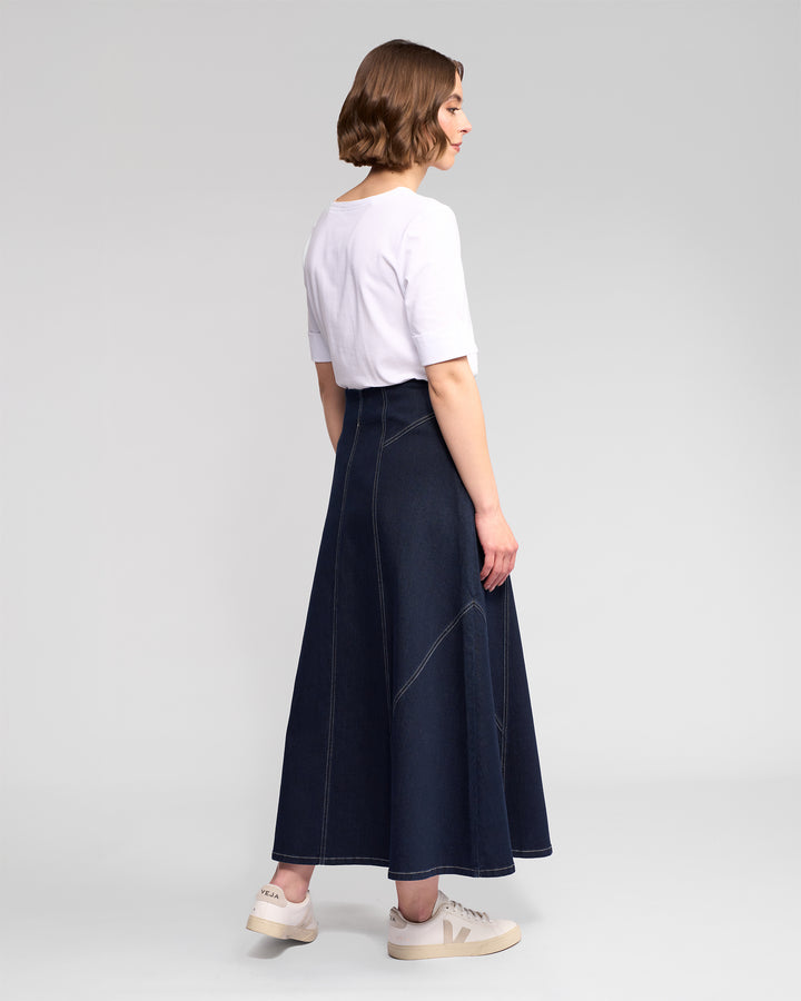 A woman faces away, wearing a white short-sleeve top, white sneakers, and the Vassalli Akari Skirt in Ink Denim with contrast white stitching. The background is plain and light gray.