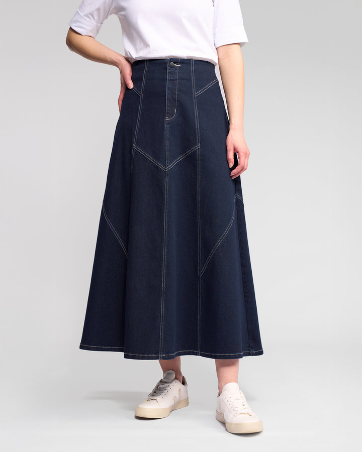 A person wears a white short-sleeve top, Vassalli Akari Skirt in ink denim with contrast white stitching, and white sneakers, standing against a plain light gray background.