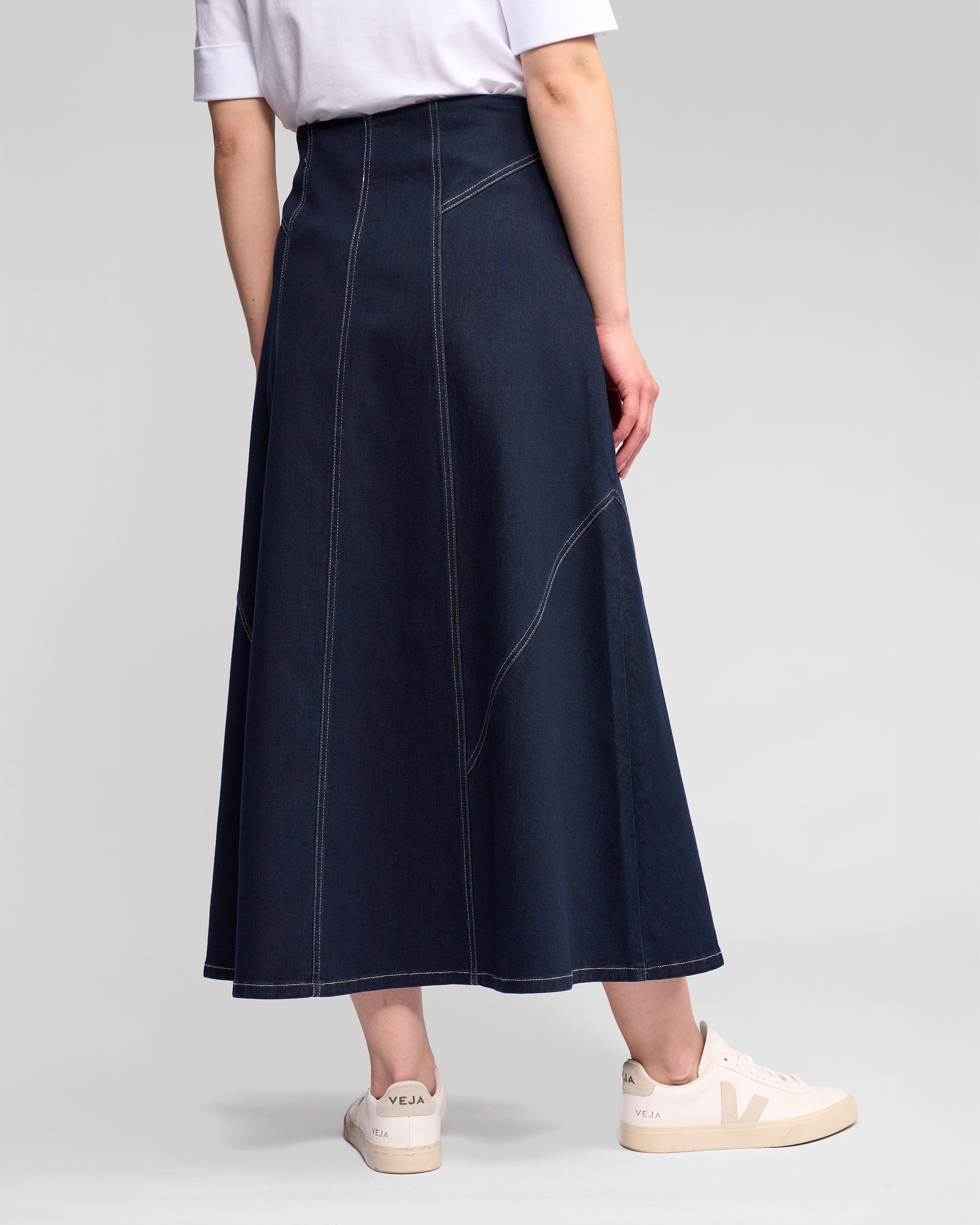 A person stands against a plain light background, shown from the back, wearing a white T-shirt, the Vassalli Akari Skirt in ink denim with contrast white stitching and visible seams, and white Veja sneakers.