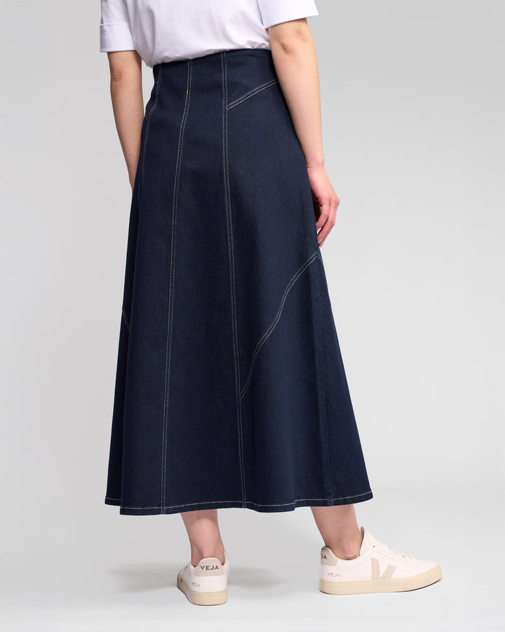 A person wearing a white T-shirt, the Vassalli Akari Skirt in ink denim, and white Veja sneakers stands against a plain light gray background.