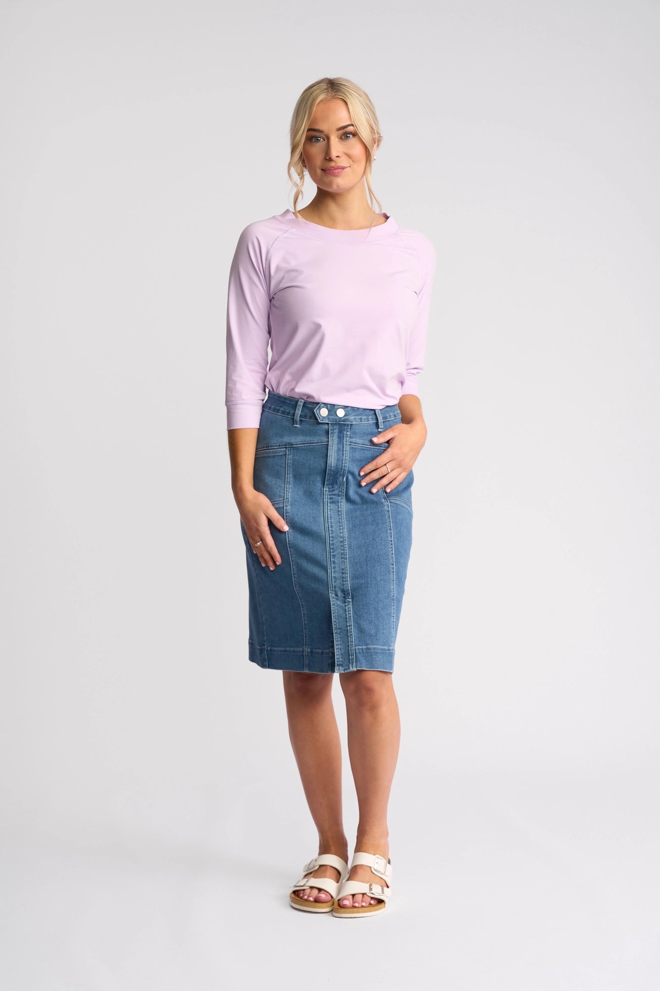 A woman stands against a plain background, wearing a light purple long-sleeve top, the Vassalli Vespa Skirt in Blue Hue Denim, and white sandals. Her blonde hair is tied back and she looks at the camera with a slight smile.
