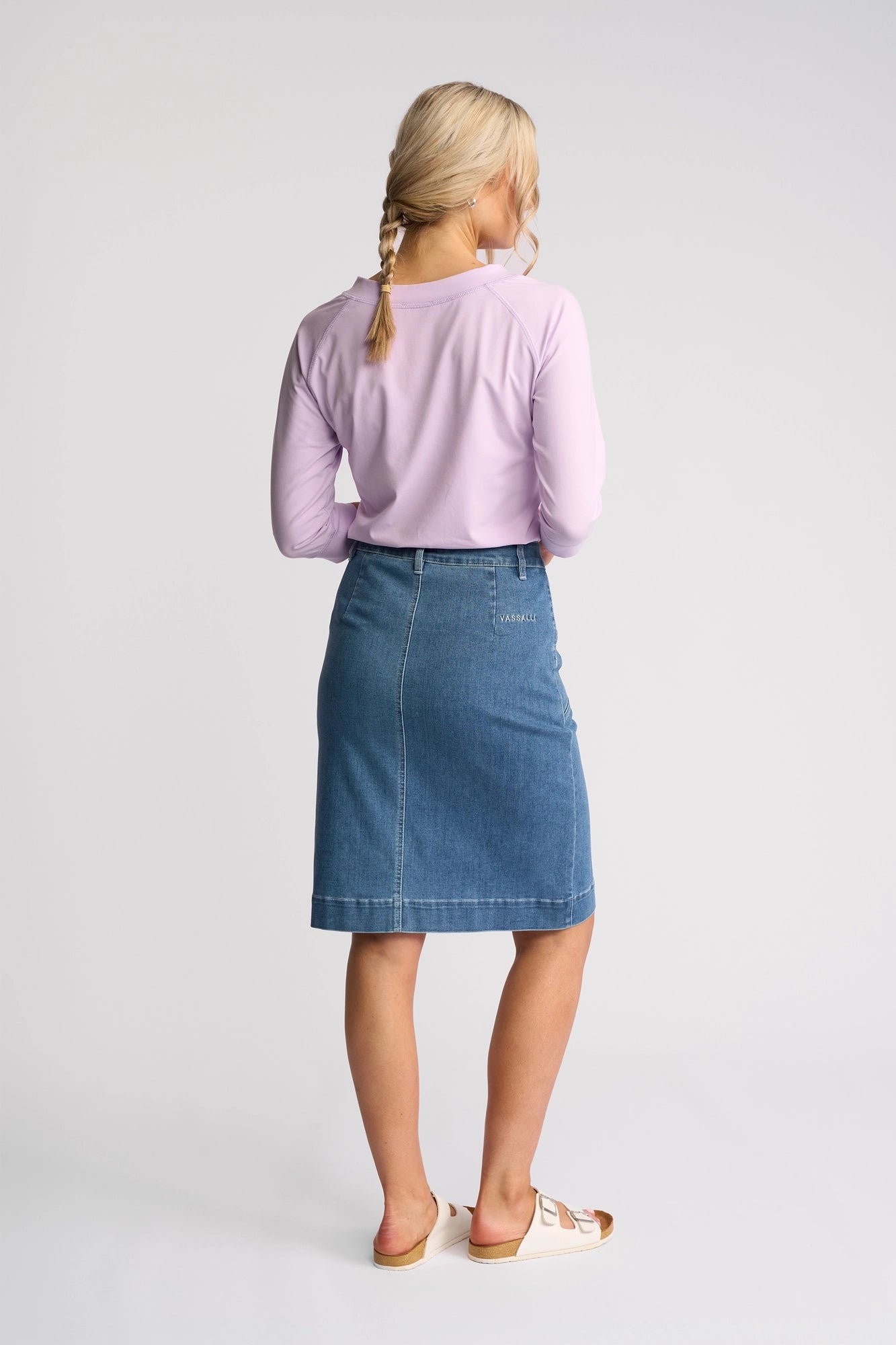 A woman with blonde hair in braids, wearing a light purple long-sleeve shirt, the Vassalli Vespa Skirt in Blue Hue Denim crafted from stretch denim, and white sandals stands facing away against a plain white background.