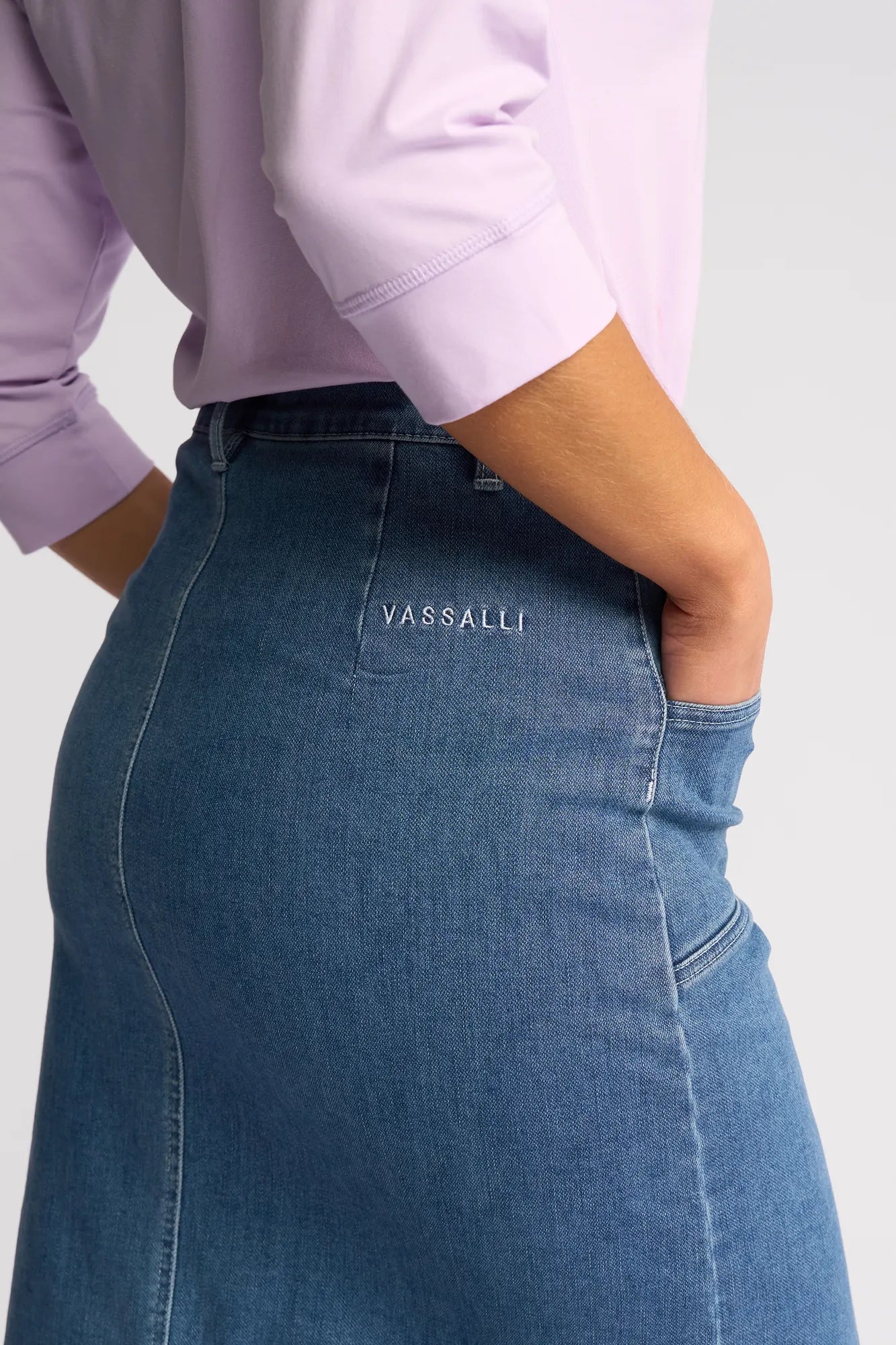 A person wearing a light purple long-sleeve shirt and the Vassalli Vespa Skirt in Blue Hue Denim, featuring stretch denim fabric and ‘VASSALLI’ embroidered on the pocket, stands with their hand in the pocket.