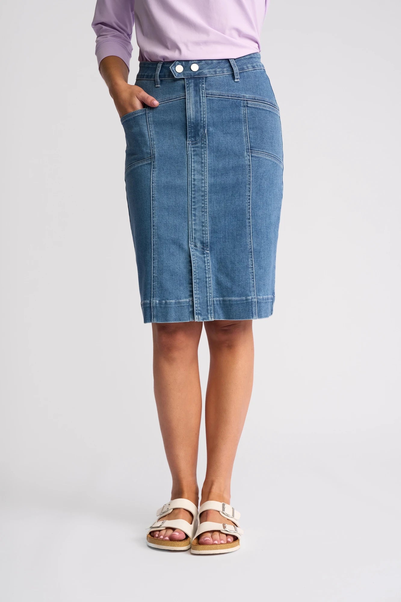 A person wearing a knee-length Vassalli Vespa Skirt - Blue Hue Denim, paired with a light purple top and white sandals, stands with one hand in a pocket against a plain background.