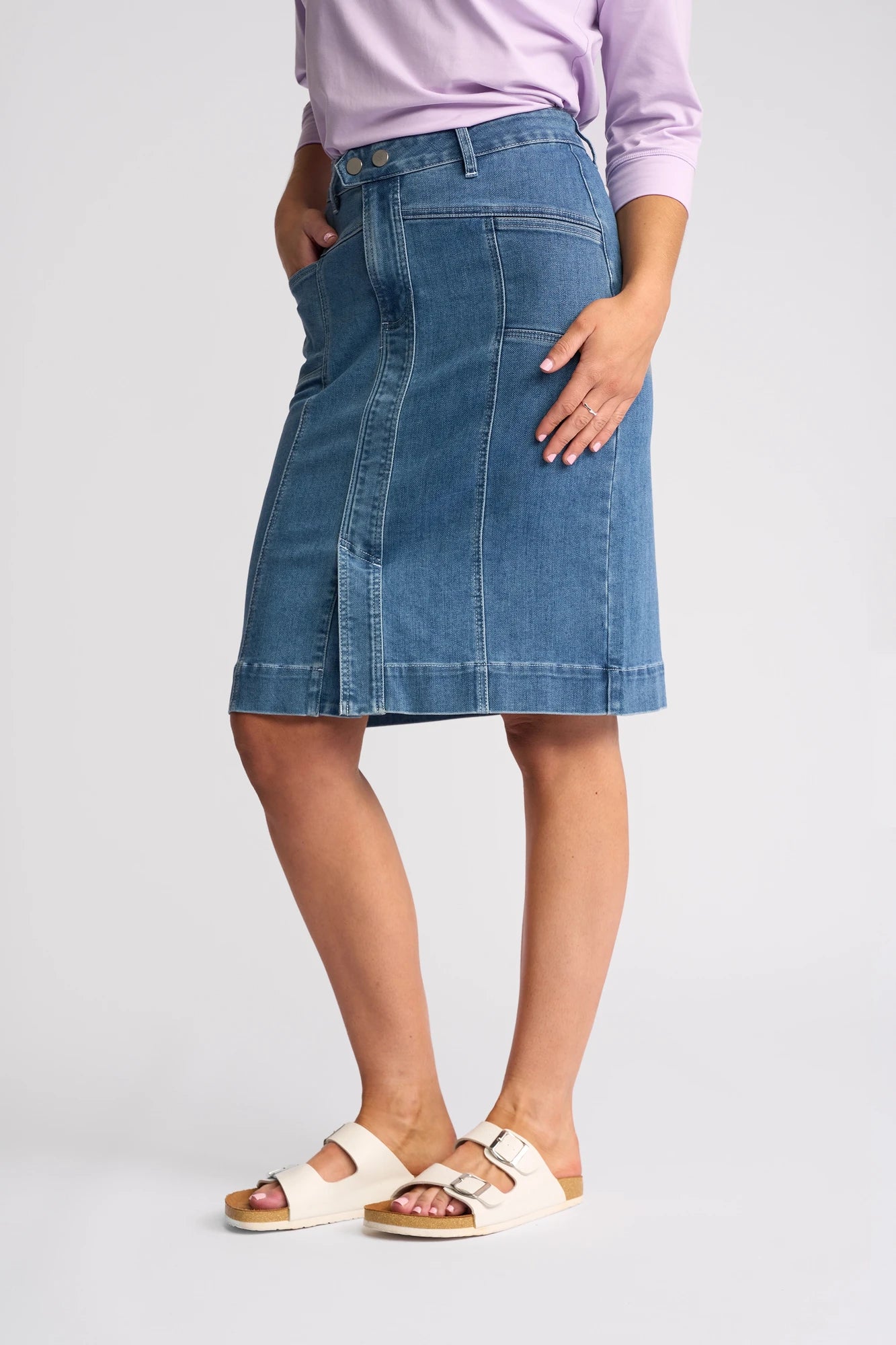 A person wearing a light purple top, the Vassalli Vespa Skirt - Blue Hue Denim featuring front stitching and a knee-length stretch fit, along with white sandals, stands against a plain light background.