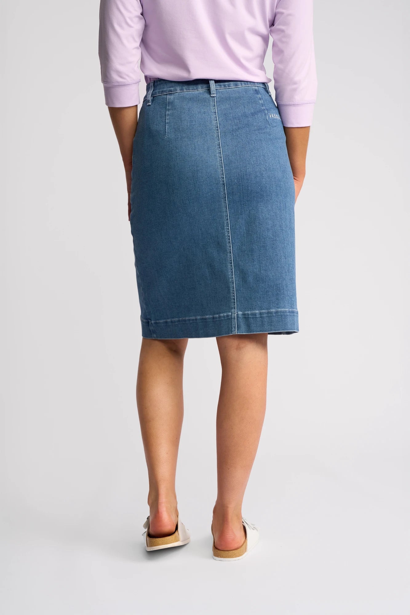 A person wearing a light purple top, the Vassalli Vespa Skirt - Blue Hue Denim—a stylish blue denim skirt from Vassalli—and white slip-on shoes stands facing away, displaying the back of the outfit against a plain background.
