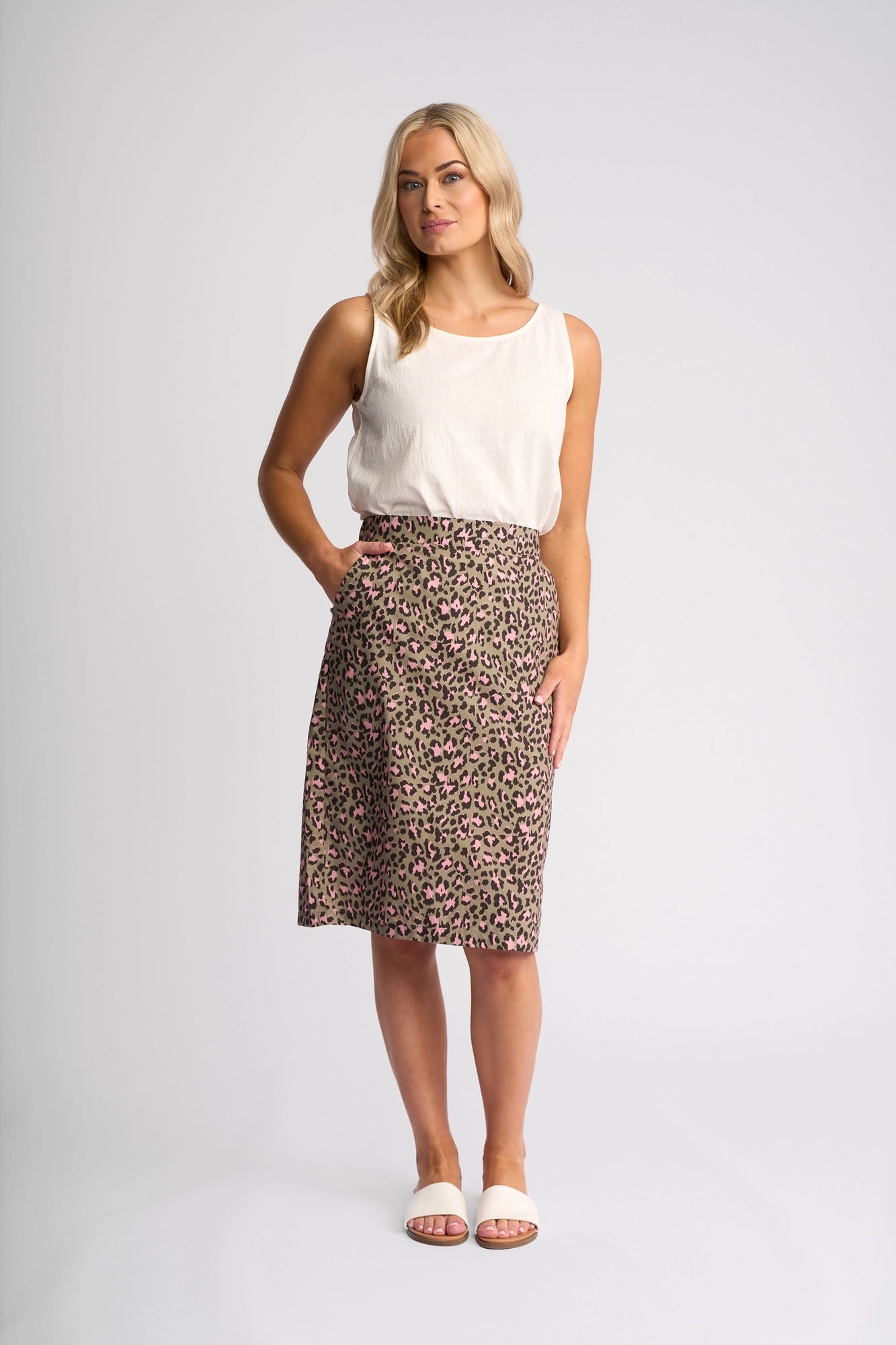 A woman with blonde hair stands against a white background, wearing a sleeveless white top, white sandals, and the Vassalli Frankie Skirt - Cub—a knee-length pink and brown leopard print skirt—with one hand in her skirt pocket.