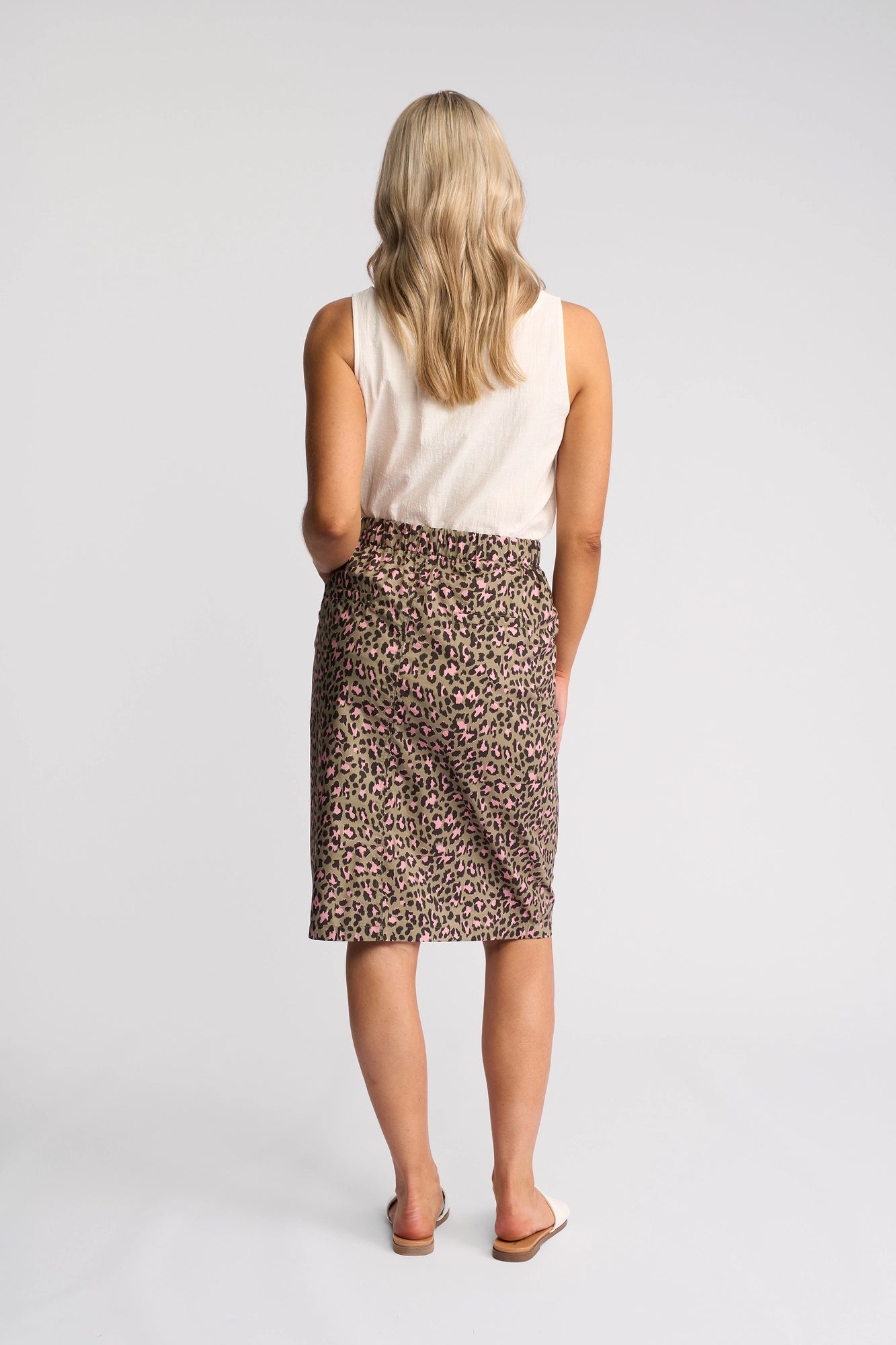 A woman with blonde wavy hair stands facing away, wearing a sleeveless white top, the Vassalli Frankie Skirt - Cub, and white slip-on sandals against a plain white background.
