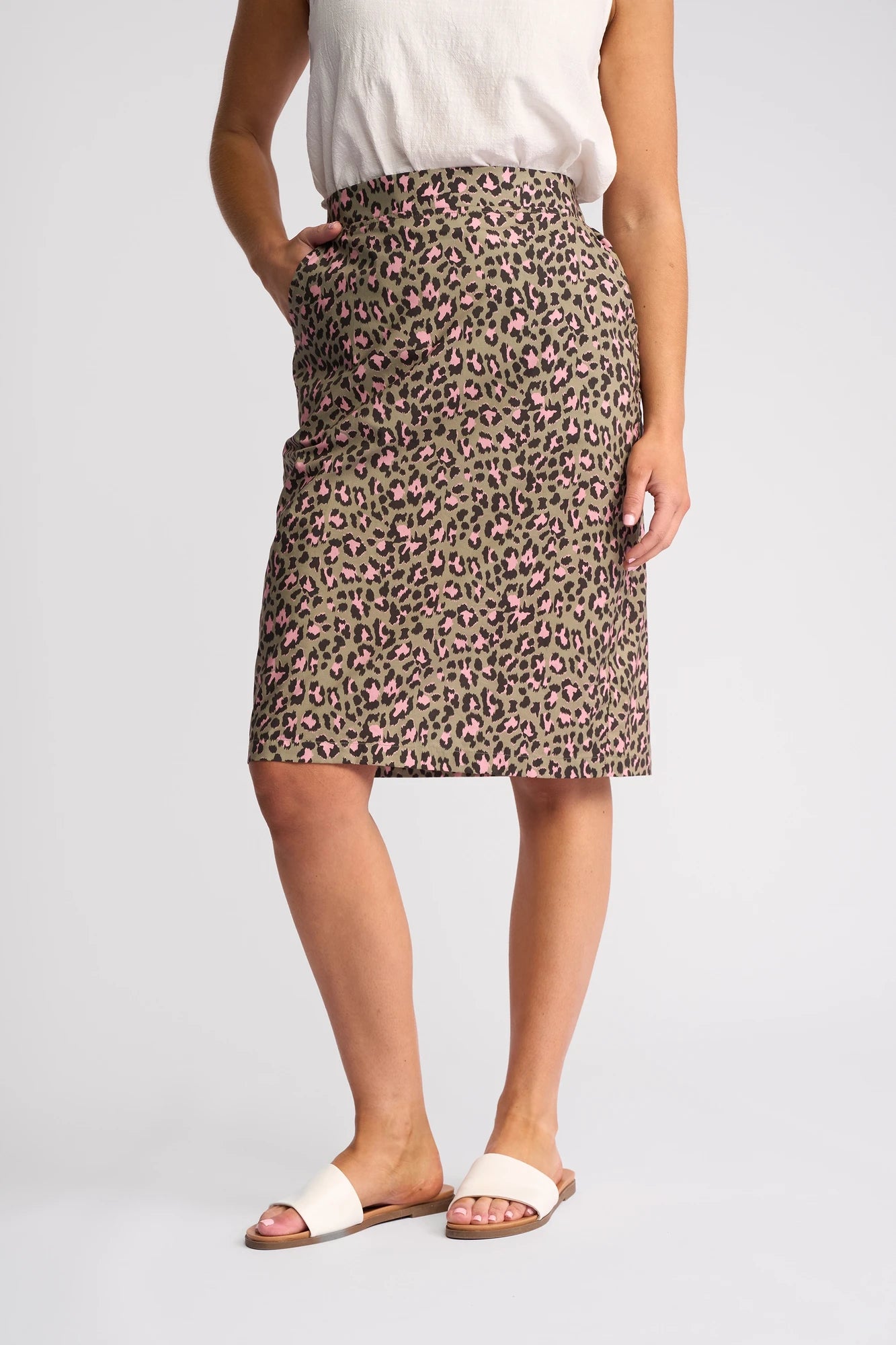 A person wearing a white sleeveless top, the Vassalli Frankie Skirt - Cub (a knee-length skirt with a leopard print and pink accents), and white slide sandals stands with their hands in the skirt pockets against a plain background.