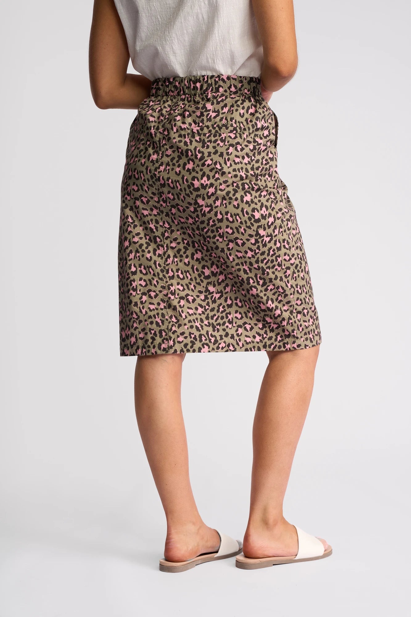 A person wearing a white top, the Vassalli Frankie Skirt - Cub—a knee-length cotton skirt featuring a leopard print with pink accents—and white slide sandals, standing against a plain light background.