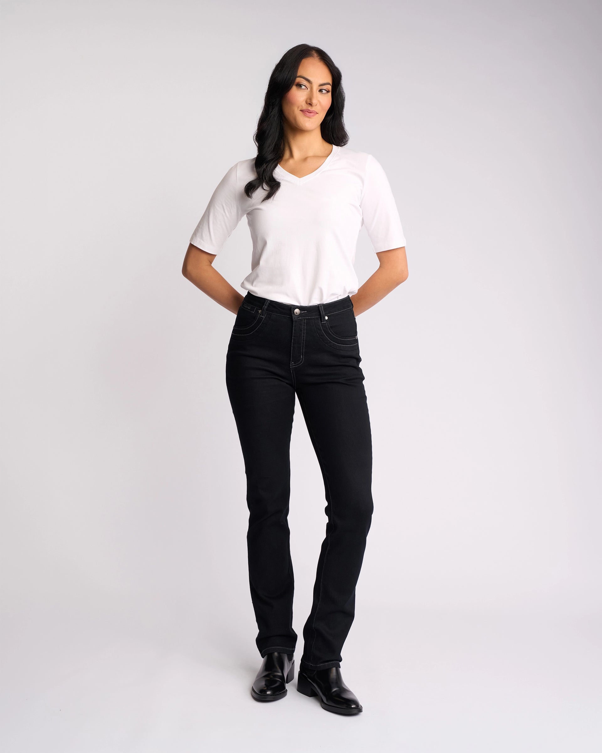 A woman with long dark hair stands against a white background, wearing Vassalli's Studio Jean in Black Denim, a white short-sleeve shirt, and black shoes. She smiles slightly with her hands on her hips.