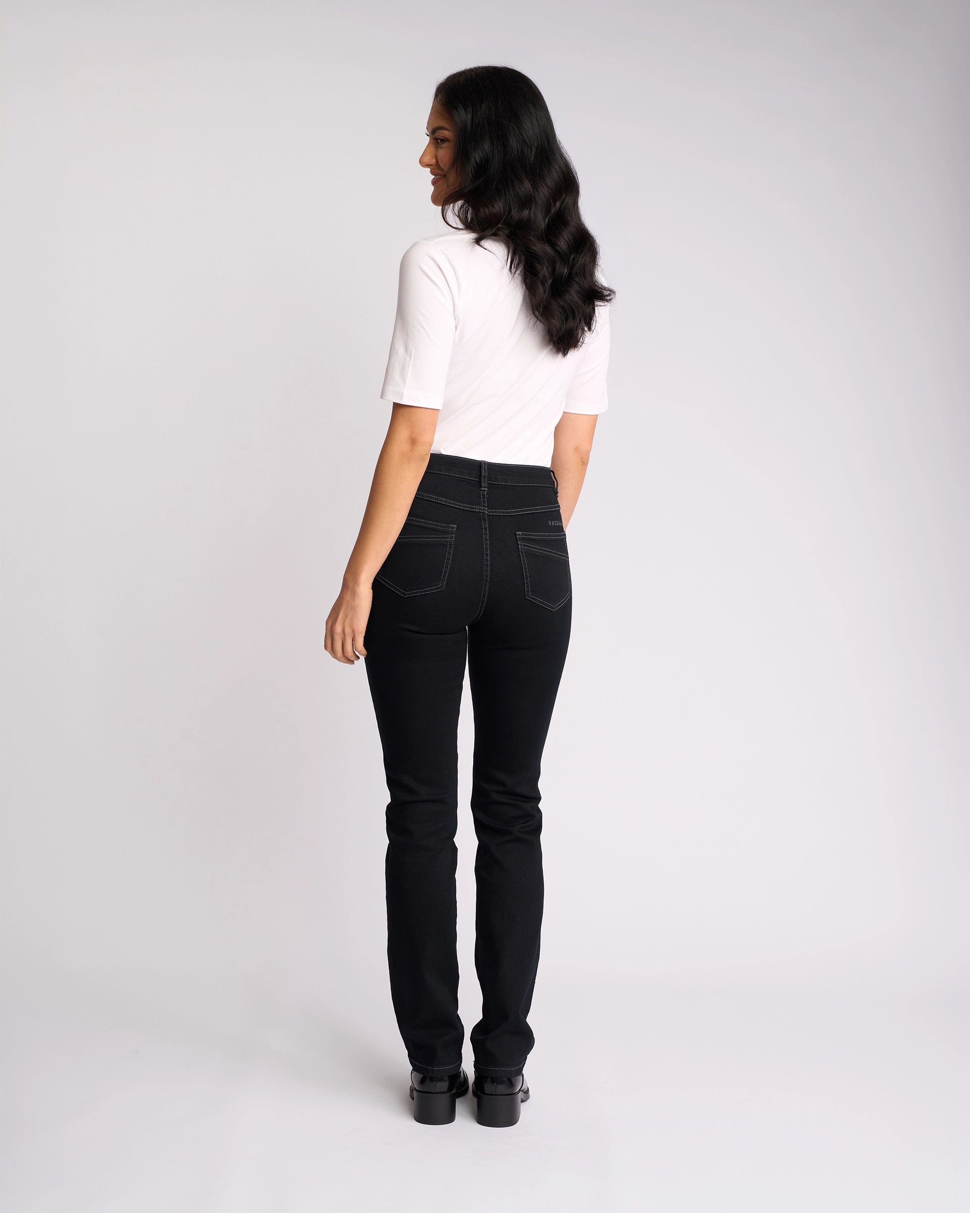 A woman with long dark hair faces away, wearing Vassalli’s Studio Jean in black denim, a white short-sleeve top, and black shoes, set against a plain light background.