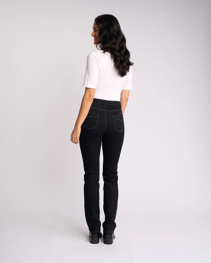 A woman with long dark hair faces away, wearing Vassalli’s Studio Jean in black denim, a white short-sleeve top, and black shoes, set against a plain light background.